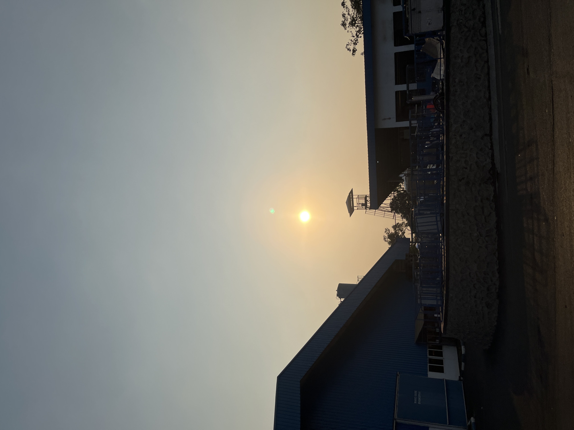 The image depicts an outdoor scene at sunset, likely in Pelang, Indonesia, characterized by a warm, hazy sky. In the foreground, a dark asphalt surface suggests a road or open area. To the left, a large, blue industrial building with a sloped roof dominates the frame. Its facade features corrugated metal siding and a section with visible windows and text reading "WAREHOUSE ENGINEERING." Adjacent to this building, a circular, low-lying structure with a stone retaining wall is visible, topped with blue railings and what appears to be industrial equipment or seating arrangements.
Further to the right, a smaller, white building with blue trim stands, partially obscured by trees. A prominent feature above this building is a tall, metal observation tower or structure with a small, roofed platform at its apex. The sun is a bright, glowing orb in the sky, casting a warm, diffused light across the scene and creating lens flare in the upper right quadrant. The overall environment appears to be an industrial or commercial complex, possibly a factory or logistics hub, set against a natural backdrop of trees. No people are visible in the image.