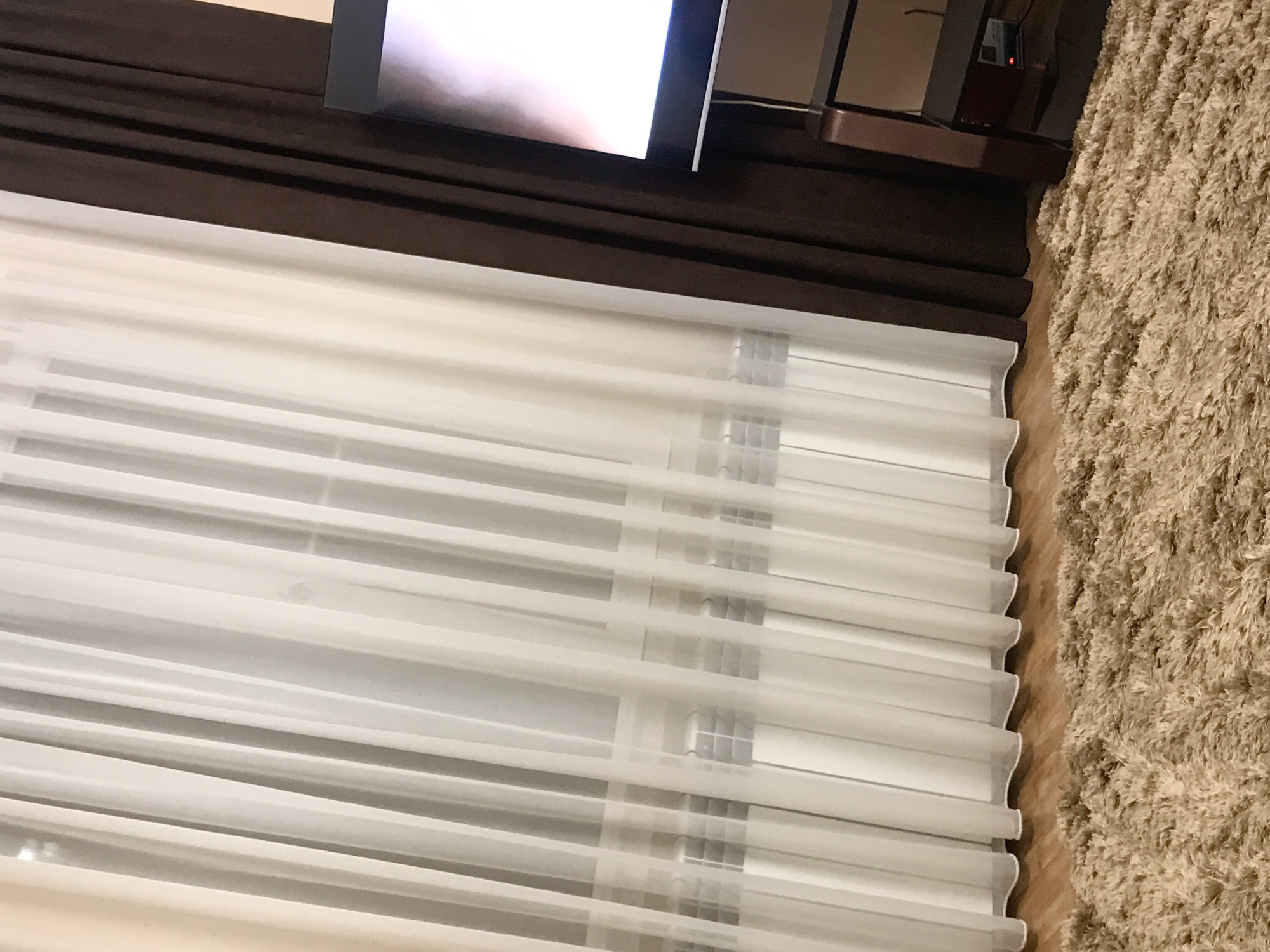 A close-up indoor shot captures white blinds partially closed, positioned beneath a dark brown curtain rod and what appears to be a television. The blinds are horizontally aligned, creating a rhythmic pattern. Adjacent to the blinds on the right is a textured, beige carpet. Based on the available information, determining the specific location within Yerevan, Armenia, is not possible.