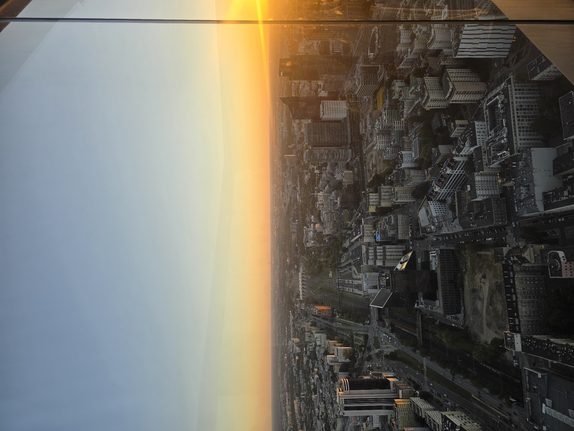 This is an aerial photograph of Warsaw, Poland, taken during sunset, likely from a high-rise building. The city stretches out to the horizon, characterized by a dense urban landscape of modern skyscrapers and older, lower-rise buildings. A prominent feature of the scene is a set of train tracks running through the middle of the city, flanked by buildings and some greenery. The sky is a gradient of soft blues and warm oranges, with the sun's bright glare visible on the right side of the frame, reflecting off a glass structure that appears to be part of the viewpoint. The overall impression is that of a bustling metropolis at the end of the day.