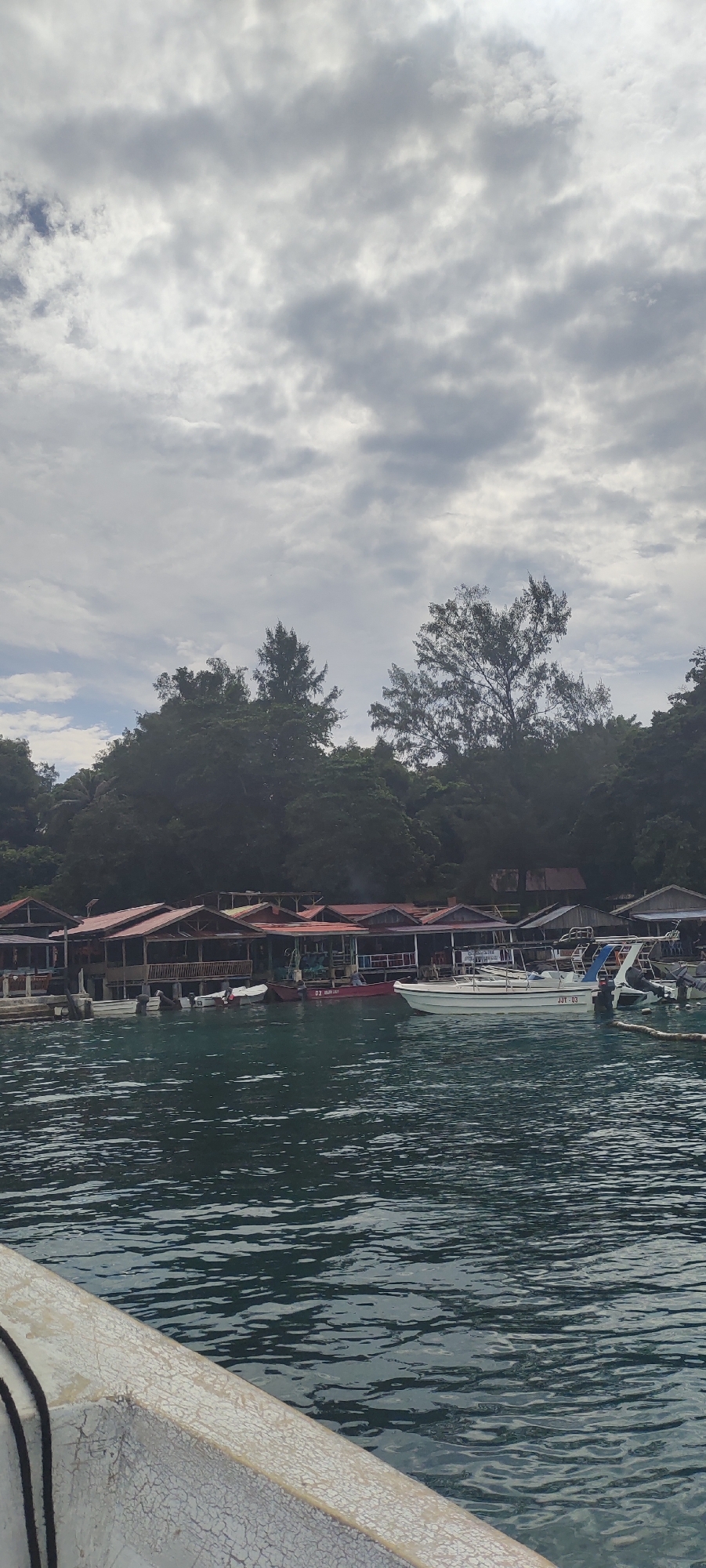 The image captures a waterfront scene with several stilted buildings featuring red roofs, likely restaurants or shops, lining the shore. Lush green trees form a dense backdrop behind the buildings. Numerous small boats and a larger white speed boat are moored in the clear turquoise water. The sky above is overcast with heavy, grey clouds, suggesting a cloudy or potentially rainy day. The foreground shows a portion of a white boat with a weathered exterior, indicating it's a vessel from which the photo was taken. The location is identified as the city of Sabang, Indonesia.