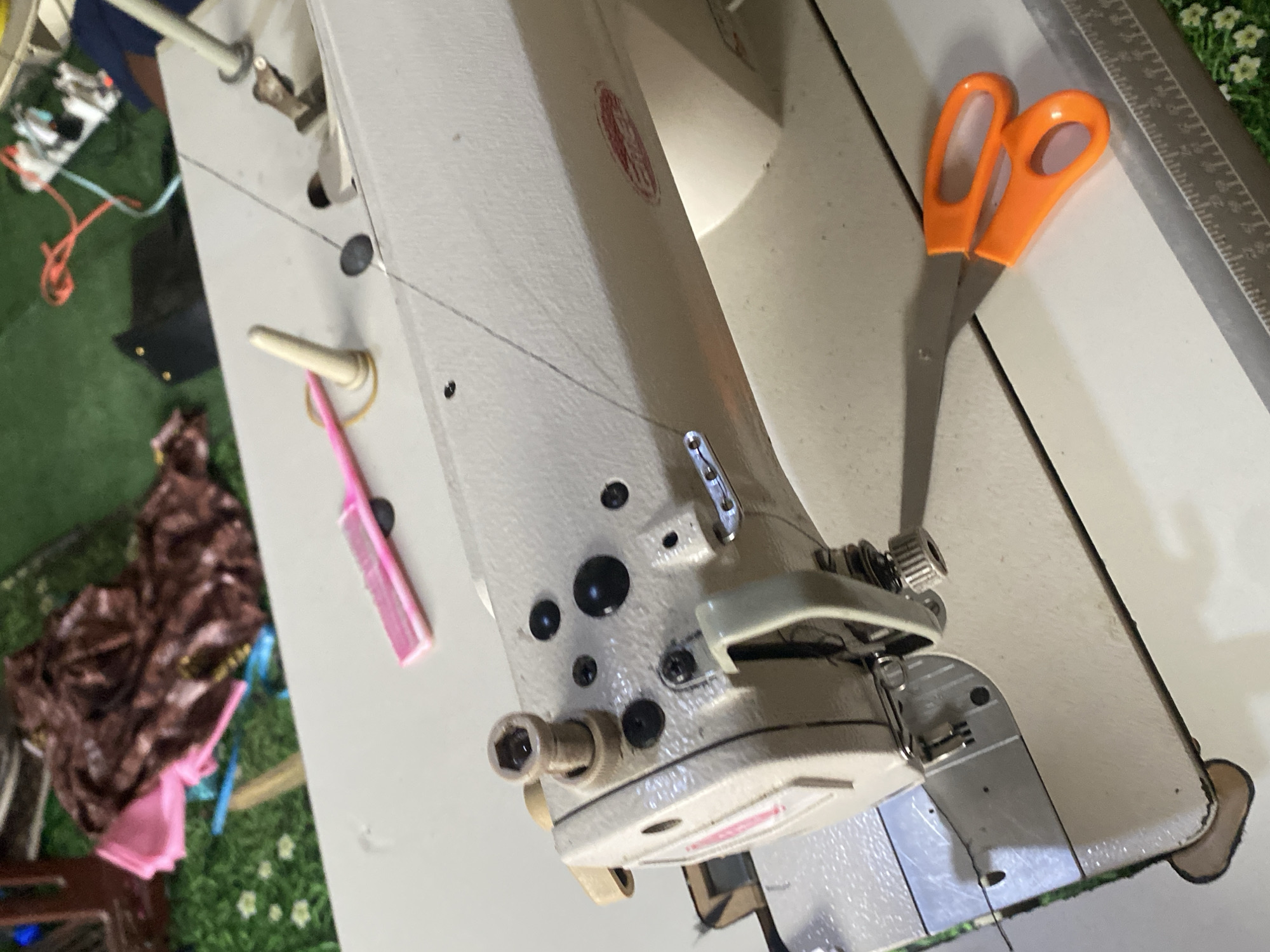 The image depicts a sewing machine with various sewing-related items on a table. The sewing machine is light-colored and appears to be in use, with a thread running through it. To the right of the sewing machine, a pair of bright orange scissors is placed. Further to the left, a pink comb and a rubber band are visible on the machine's surface. In the background, fabric scraps, including pink and brown material, are scattered on a green, artificial turf-like surface, suggesting a workspace. The overall environment seems to be an indoor setting, possibly a tailor's shop or a home sewing area. The presence of the sewing machine, fabric, and tools indicates an activity related to sewing or garment creation. There is no visible indication of the city of Jauro Yinu, Nigeria within the image itself.