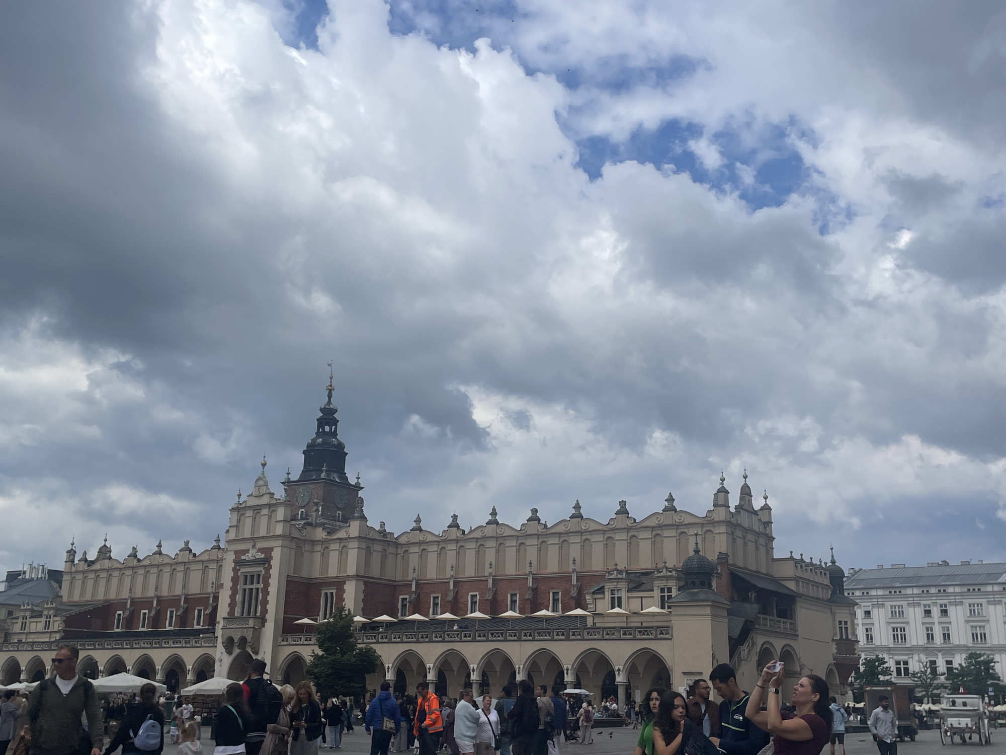 The image captures a bustling scene in Krakow, Poland, specifically at the Main Market Square (Rynek Główny). The iconic Cloth Hall (Sukiennice) dominates the center of the frame, its historic architecture featuring a long, arcaded ground floor and an ornate facade. A prominent clock tower stands tall on the Cloth Hall. The sky above is filled with dramatic, grey clouds, suggesting overcast weather. A diverse crowd of people is visible, walking, conversing, and taking photographs. Some individuals are captured mid-action, such as a woman in the foreground holding up a phone to take a picture. The square appears to be a lively public space with people enjoying their day amidst the historical surroundings.