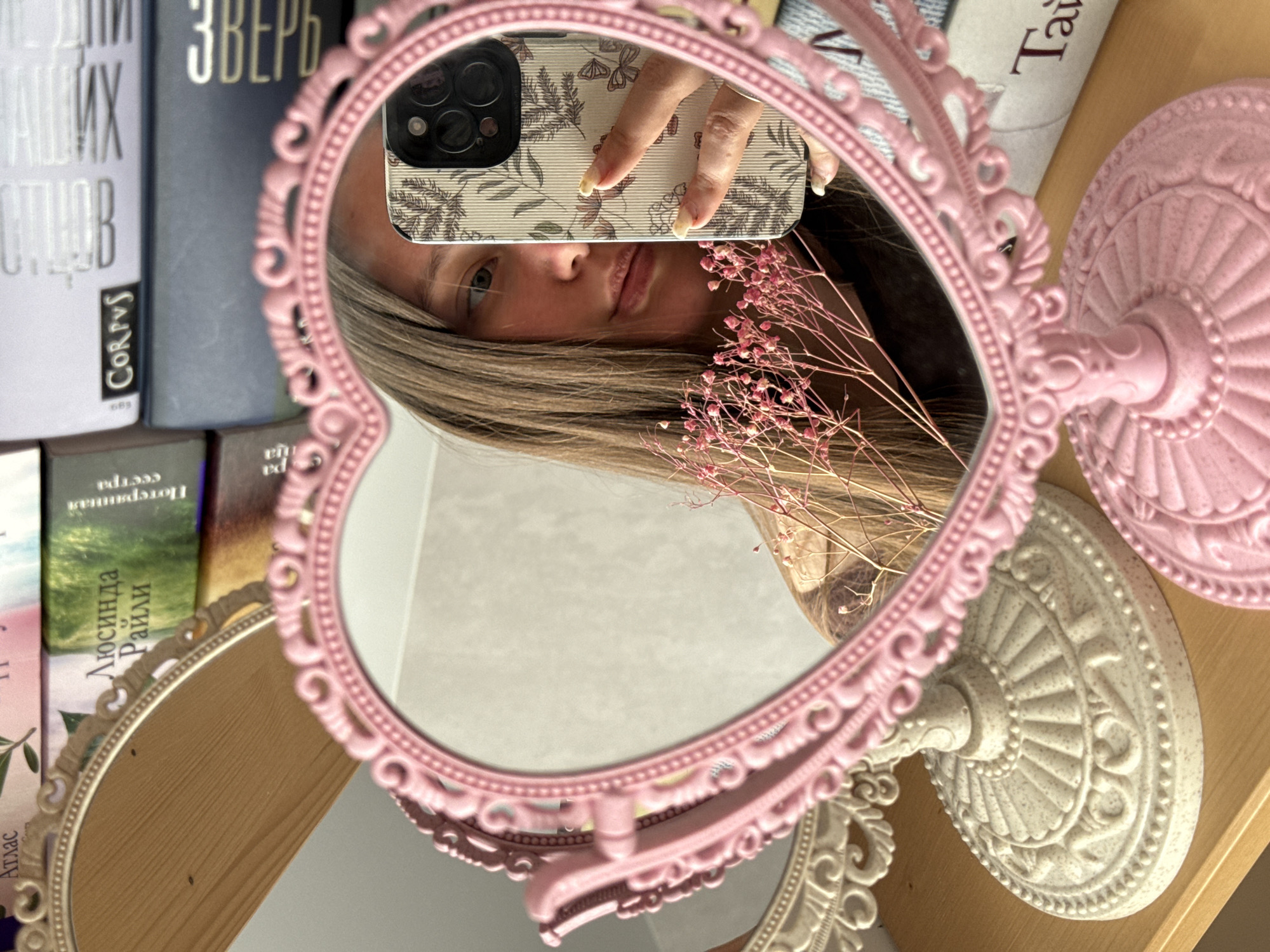 This vertical selfie is captured as a reflection in an ornate, heart-shaped pink mirror. The image itself is rotated 90 degrees counter-clockwise.
The primary subject is a person, likely female, whose face is partially visible in the mirror's reflection, showing her nose, mouth, chin, and parts of her eyes. Her long, light brown hair frames her face. She is holding a smartphone, visible in the upper part of the reflection, with her hand positioned near the phone's triple camera lenses. The phone case features a delicate pattern of dark leaves and butterflies on a light, possibly cream or beige, background. A sprig of dried pink gypsophila-like flowers rests on the wooden surface next to the reflected face.
The pink mirror, with its decorative, rococo-style frame and fluted stand, is the central element of the composition. It stands on a light-colored wooden shelf or table. In the background, behind the mirror, are several books stacked both horizontally and vertically. Visible text on the book spines includes "ЗВЕРЬ" (ZVER, meaning BEAST) on a dark blue book, "CORPUS" and "683" on a light purple book, "Люсинда Райли" (Lucinda Riley) and "Потерянная Сестра" (The Missing Sister) on another, and "Атлас" (Atlas) on a spine at the bottom. Another partially visible book shows "Тай" (Tay).
A second, larger oval mirror with a cream-colored ornate frame is partially visible in the lower left corner, reflecting an indistinct part of the room. The lighting is soft and appears to be natural, casting gentle shadows. The overall mood is decorative and intimate, centered around the act of taking a mirror selfie amidst personal belongings.
