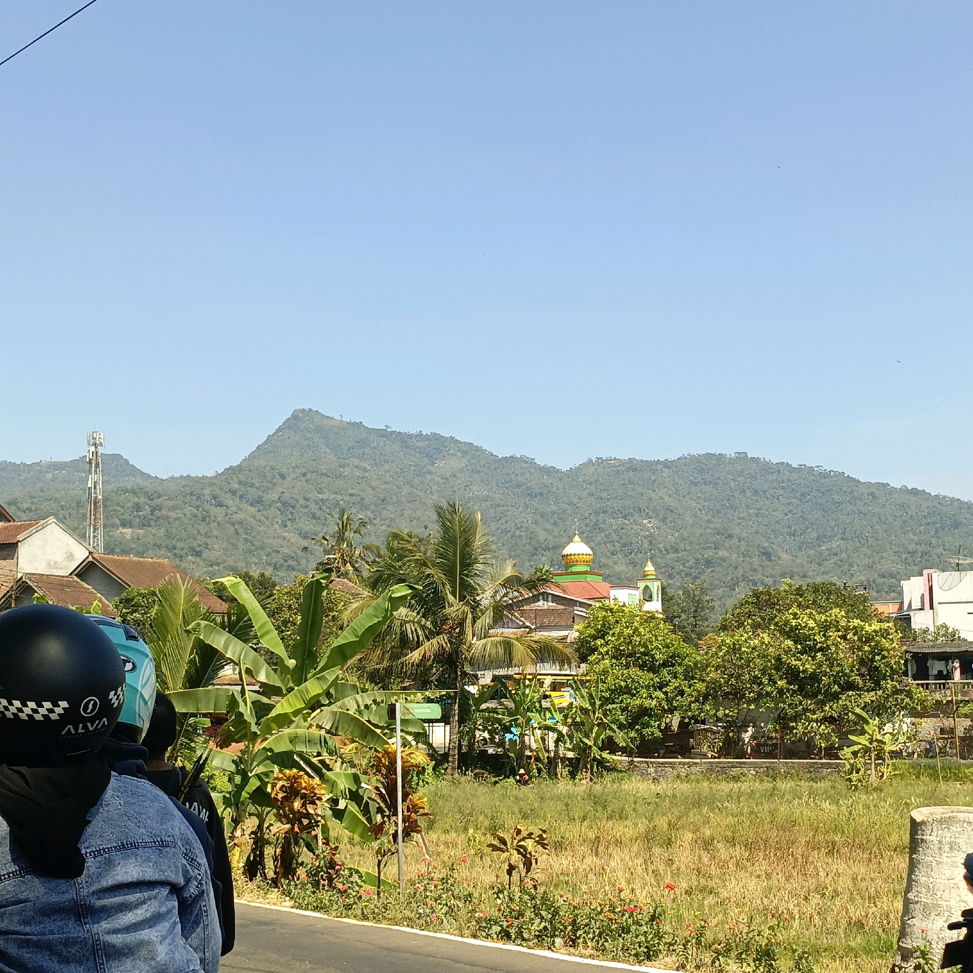 In Rowoboni, Indonesia, a sunny day displays a panoramic view of lush green hills in the background, topped by a clear blue sky. In the mid-ground, a mosque with a golden dome stands out, surrounded by trees and buildings. A telecommunications tower is visible to the left, adding a modern element to the rural landscape. In the foreground, three people on a motorcycle are seen from behind, their helmets indicating they are traveling. The motorcycle is on a paved road, with a field of dry grass and scattered flowers to the right. The scene captures a blend of natural beauty, religious architecture, and everyday life in Indonesia.