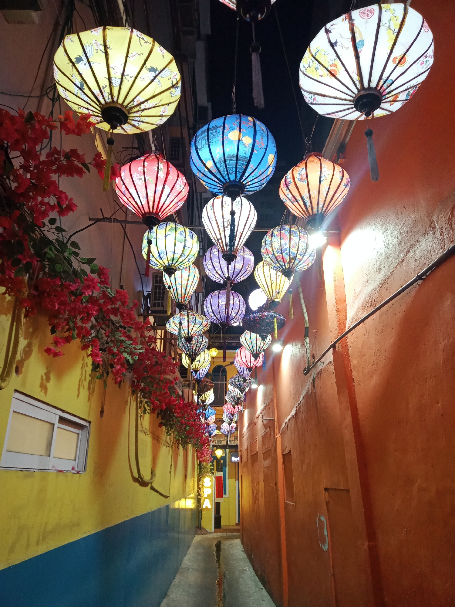 This image captures a narrow alleyway in Nha Trang, Vietnam, illuminated by a vibrant array of hanging lanterns. The walls are painted in contrasting colors; yellow and blue on one side, and orange on the other. Red bougainvillea flowers cascade from the yellow wall, adding a natural element to the urban scene. The lanterns are of varying colors and patterns, casting a warm glow down the alley. The narrow passageway leads toward a brightly lit area in the distance, suggesting a bustling street or entrance. The alley is devoid of people, emphasizing the architectural details and the festive atmosphere created by the lanterns.