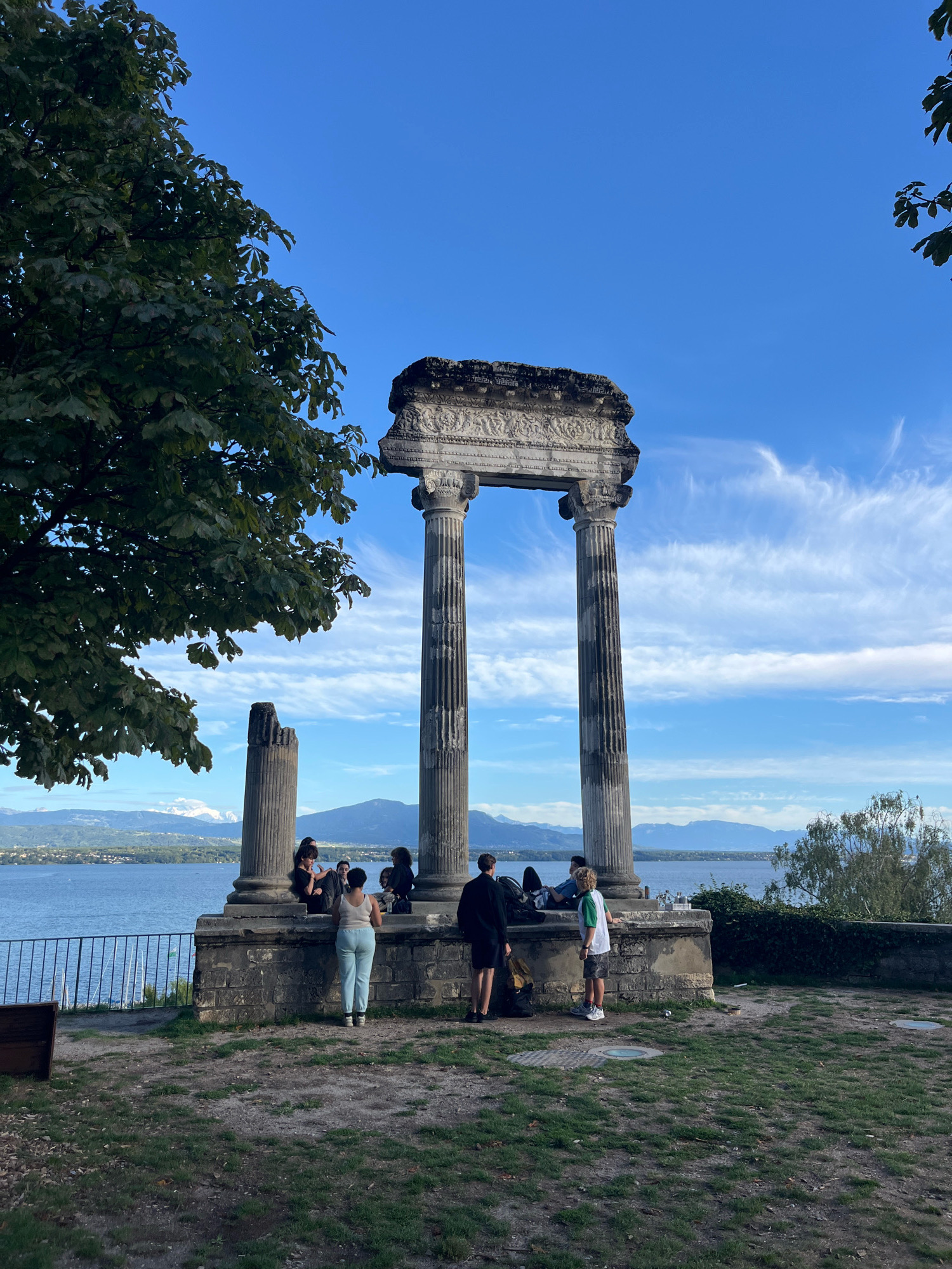 In Nyon, Switzerland, a group of individuals is gathered near ancient ruins overlooking a body of water. The ruins consist of three large stone columns, two of which support a heavily ornamented entablature, suggesting a classical architectural style. A partial, broken column stands to the left of the main structure.
Several people are visible in the foreground and midground. On the left, a person wearing a light-colored tank top and light blue pants stands with their back to the camera. They are facing a group of people seated and standing among the ruins. To the right of this individual, a person in dark clothing is also facing the ruins, with a backpack nearby. Further to the right, a young person in a white and green shirt and shorts stands, also facing the ruins. Another person in dark clothing stands to the far right, facing away from the camera.
The ruins are situated on a raised platform with stone walls and a grassy area surrounds it. Beyond the ruins, a vast body of water, likely Lake Geneva, stretches towards a distant shoreline with mountains. The sky is a vibrant blue with scattered white clouds, some wispy and others more defined. Trees with green foliage are present in the foreground and to the right of the scene. A metal railing runs along the edge of the platform overlooking the water. The overall impression is a scenic overlook with historical remnants and people enjoying the view.