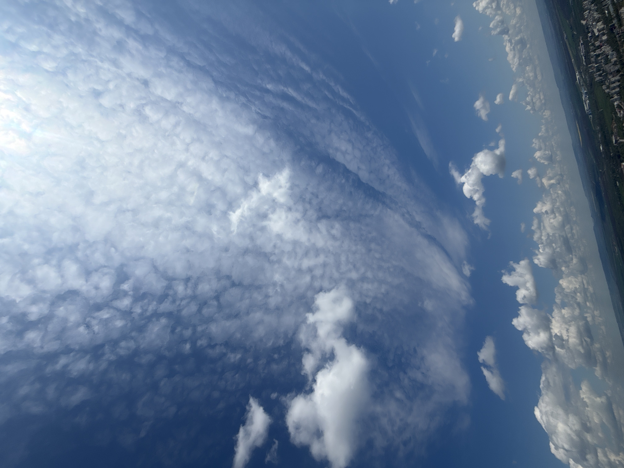 The image captures a vast expanse of a partially cloudy sky, seen from what appears to be an elevated perspective over a landscape. When properly oriented (rotated 90 degrees clockwise), a distant cityscape and green areas are visible along the horizon at the bottom right.
The majority of the sky is dominated by a sweeping band of white and light gray altocumulus clouds, characterized by their small, lumpy, and patchy appearance, often arranged in waves or rows. These clouds stretch diagonally across the sky from the upper left, becoming denser and brighter towards the far left, suggesting the direction of the sun or a heavily cloud-covered area.
Towards the right and lower sections of the sky, the altocumulus clouds are less dense, interspersed with clearer patches of deep blue sky. Several distinct, fluffier cumulus clouds are also present in this area, particularly above the horizon, adding depth and contrast to the scene. The overall impression is one of a dynamic and layered sky.
The time of day is clearly daytime, with ample sunlight illuminating the clouds. There are no people or specific objects discernible beyond the natural elements of the sky and the distant, indistinct urban and natural landscape. No activity or event is taking place, and no visible text is present in the image. The location cues are limited to the generic cityscape and green fields on the distant horizon, consistent with the given context of Shumen, Bulgaria.