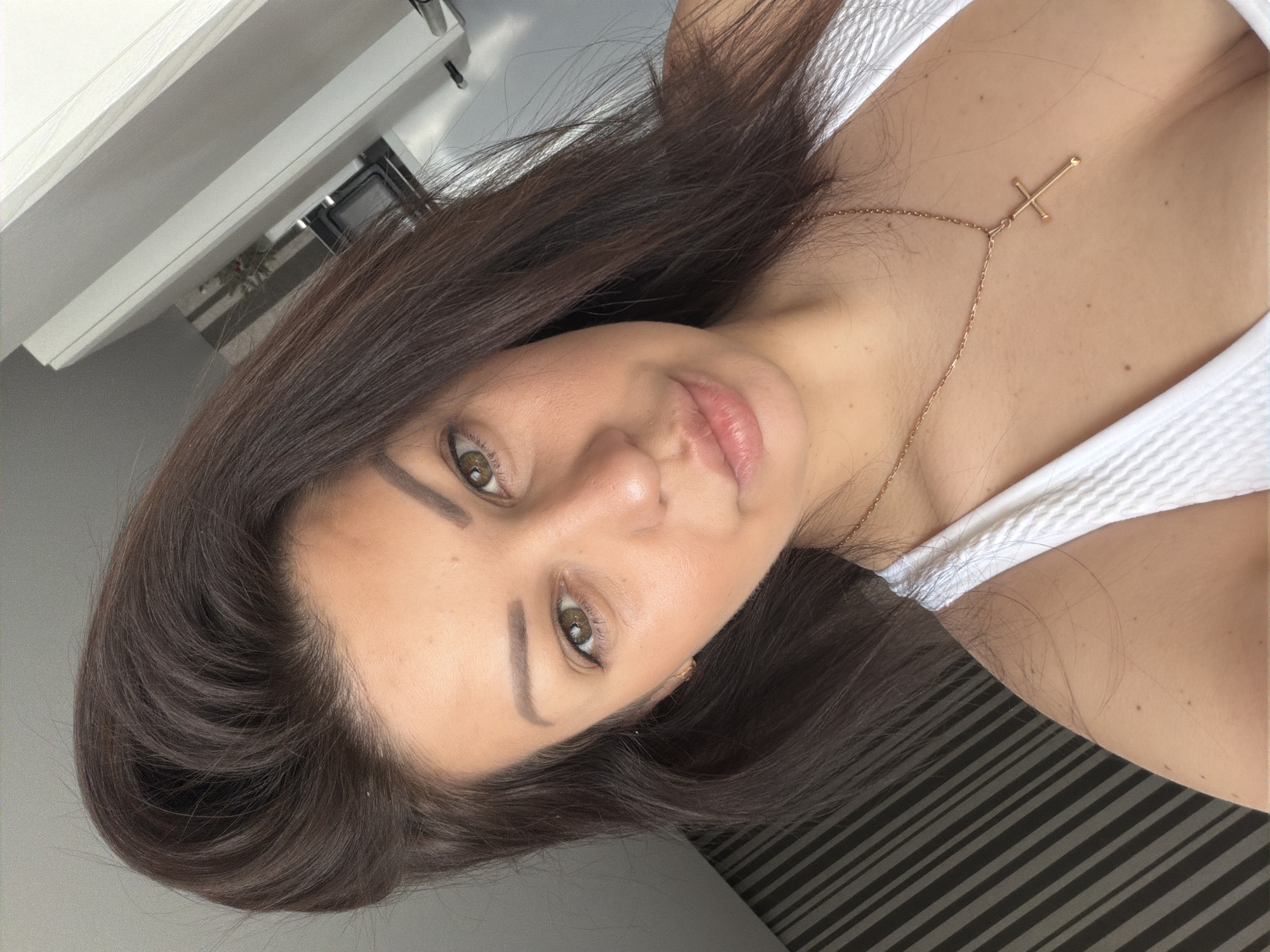 This image is a close-up selfie portrait of an adult female, likely in her 20s or 30s, taken indoors in Sochi, Russia. She has dark brown hair styled with frontal volume, hazel eyes looking directly at the camera, and visibly shaped eyebrows. Her skin complexion is fair with warm undertones, and her lips are slightly parted. She is wearing a white, textured tank top or camisole that reveals part of her upper chest. A gold-colored chain necklace with a small, plain cross pendant is visible around her neck, resting on her décolletage. Multiple small moles are present on her visible skin, particularly on her right shoulder and upper chest. The background on the left features a dark surface with diagonal light-colored stripes, consistent with blinds or wallpaper. The background on the right and top consists of light-colored, possibly white, interior surfaces, including what appears to be part of a microwave oven or kitchen appliance, indicating a domestic setting. The scene is brightly lit, likely by natural light.