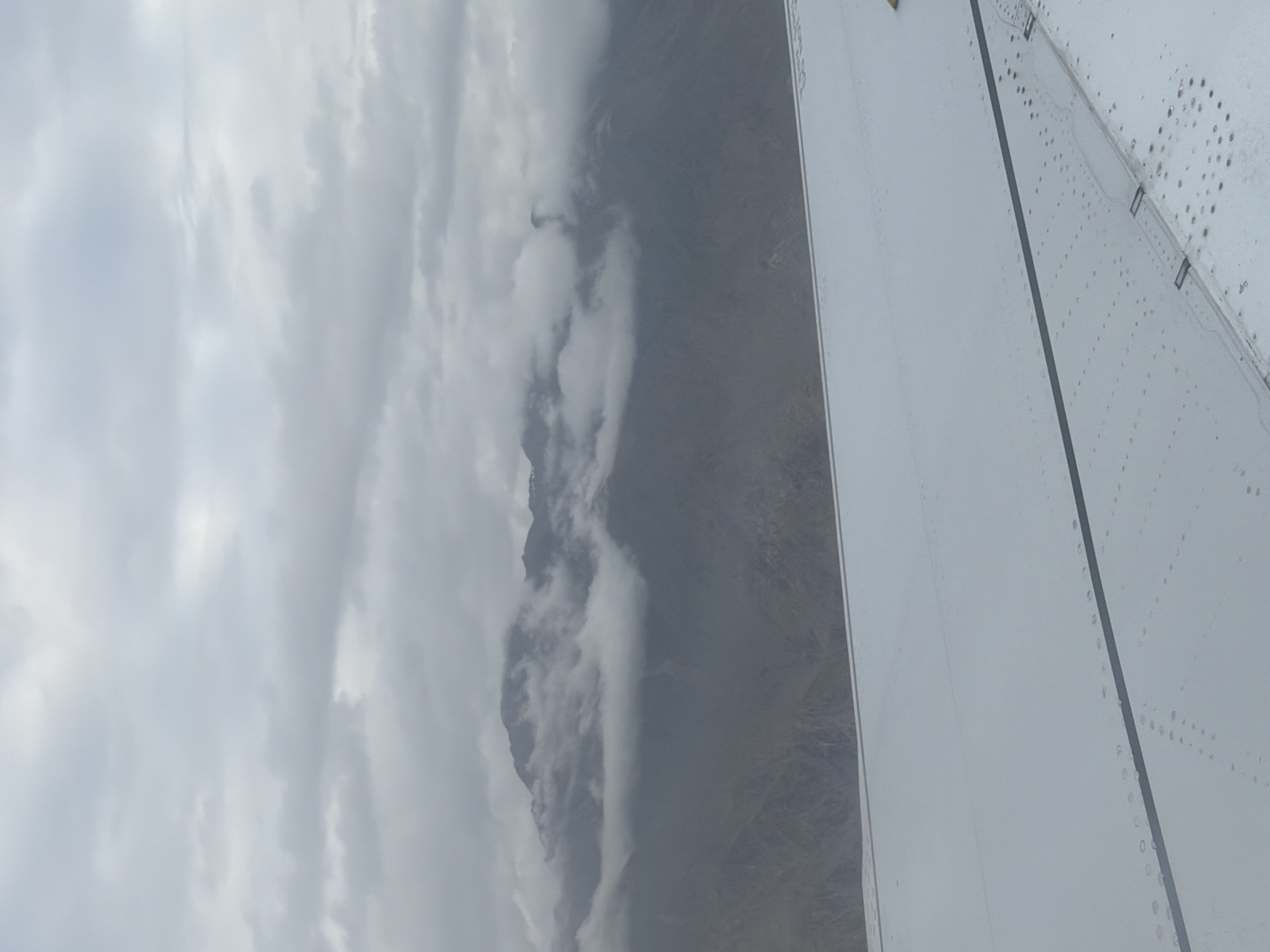 This is an aerial photograph of mountains in the city of Leh, India, viewed from an airplane window. The sky is overcast with thick, gray clouds. The mountains are rugged and appear to be covered in sparse vegetation. Clouds are obscuring the upper portions of the mountains. The foreground of the image shows the white, metallic surface of an airplane wing, with visible rivets and seams. The wing occupies the lower half of the frame, and a dark line runs horizontally across it.