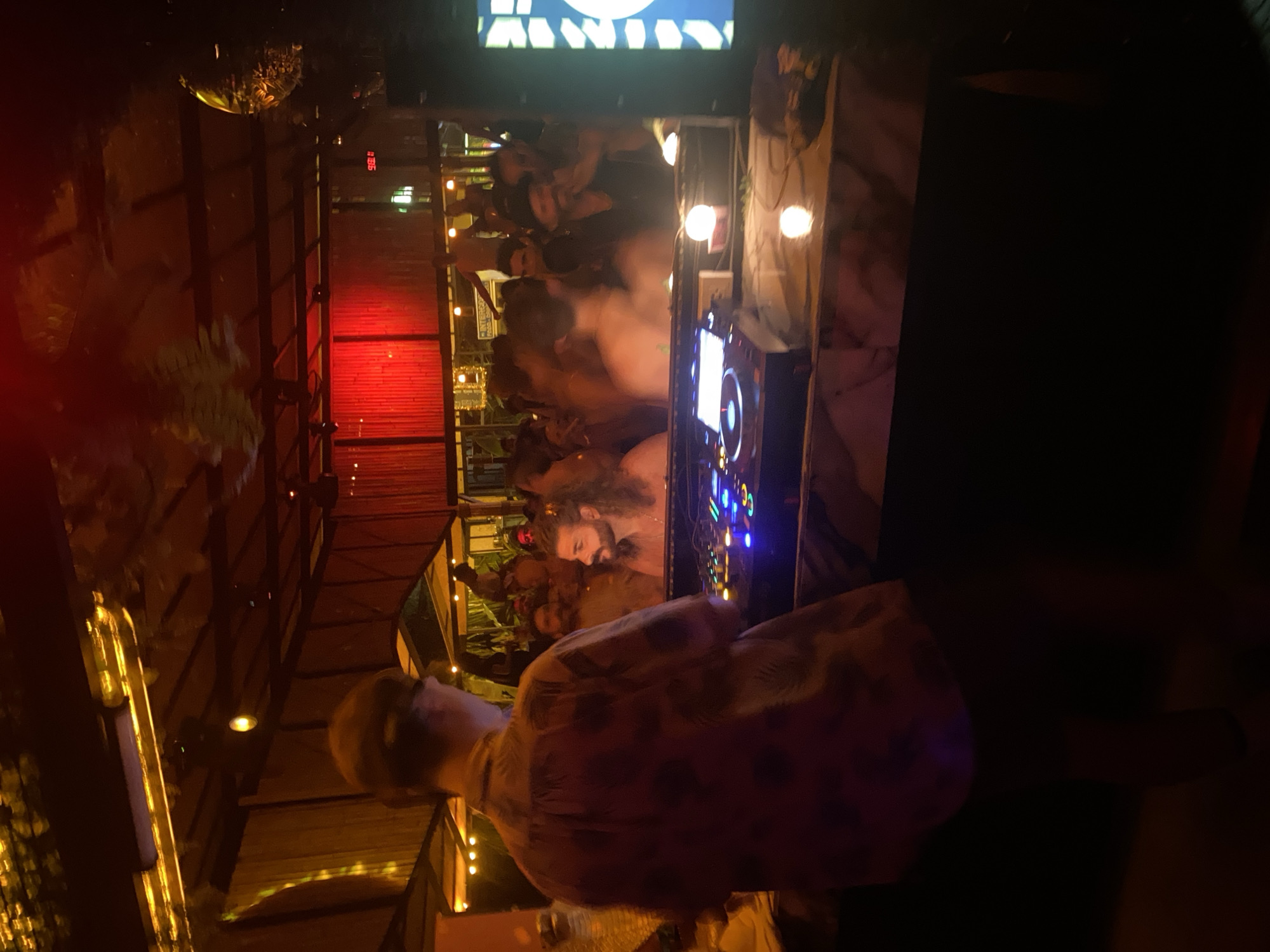 The image depicts a nighttime scene in Baan Hin Kong, Thailand, seemingly within a crowded entertainment venue. The focus is on a DJ console with a person operating it, evident from the visible controls and illuminated display. Beyond the console, a multitude of blurred figures suggest a gathering of people, possibly dancing or socializing. The environment is dimly lit with a mix of artificial lights, including some red lighting elements. There is also an overhead display screen showing possibly a logo or a visual. The structure of the venue appears to be open-air, with posts, beams, and some foliage visible in the background.