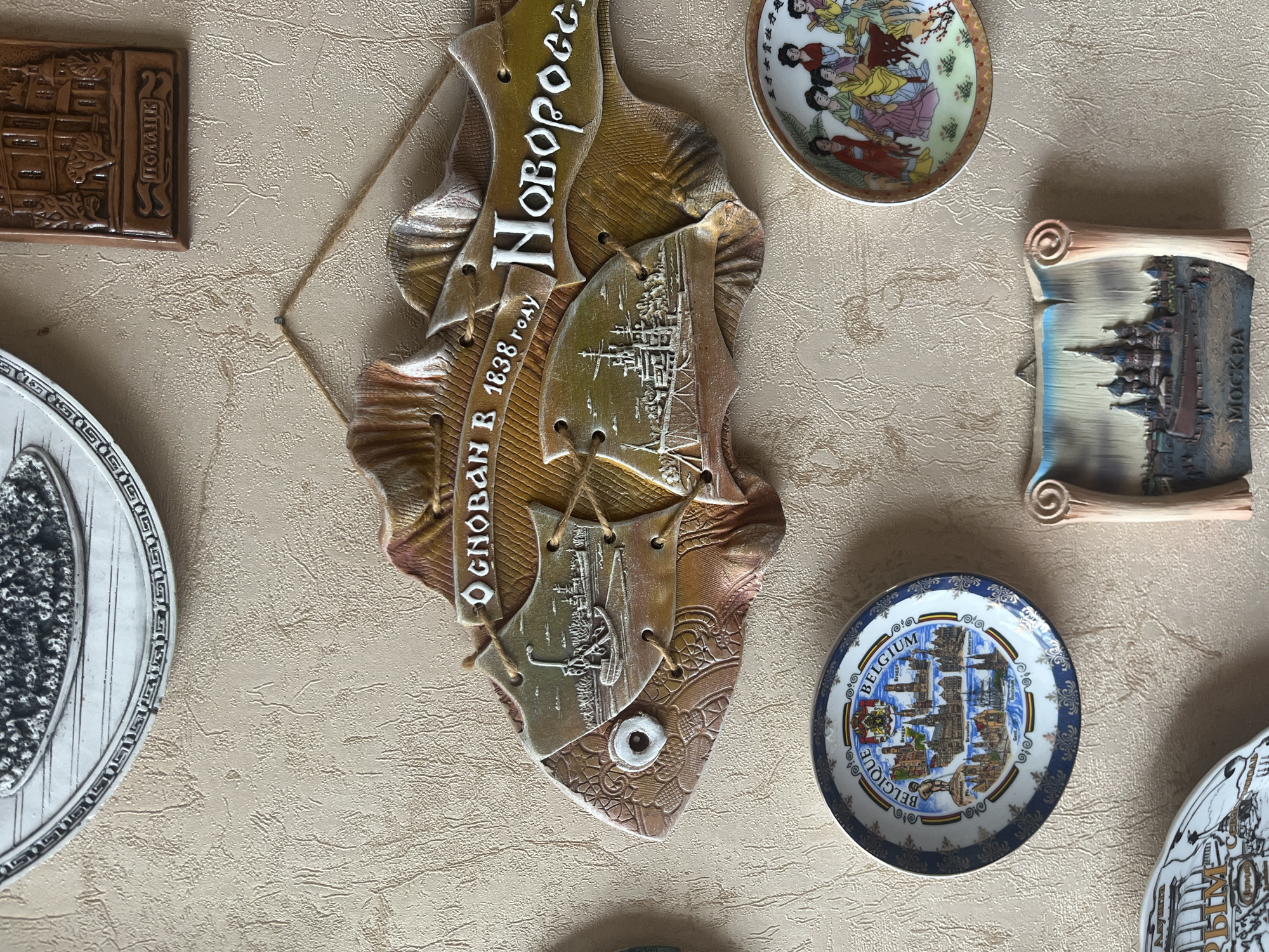 The image displays a collection of decorative plates and wall hangings on a textured, light-colored wall. In the center, a large, fish-shaped plaque features embossed imagery and text. The text on the plaque reads "ОСНОВАН В 1638 ГОДУ" (Founded in 1638) and "Новоросс" (Novoross). The left side of the fish plaque depicts a sailing ship, and the right side shows a building with towers.
Below the fish plaque, a round decorative plate with a blue and white patterned rim showcases an illustration of cityscapes and landmarks labeled "BELGIQUE BELGIUM" and names of cities like "Bruges," "Brussels," "Antwerpen," "Gent," and "Oostende." To the right of the Belgium plate, a smaller round plate features an illustration of several women in traditional Asian attire.
Further down, another decorative wall hanging, shaped like a scroll, displays a depiction of the Saint Basil's Cathedral in Moscow, with the word "МОСКВА" (Moscow) printed at its base.
In the upper right corner of the image, a small, rectangular brown plaque is visible, bearing an embossed image of a building and the word "ПОЛОЦК" (Polotsk). The uppermost item in the image is a portion of a white decorative plate with a dark, intricate pattern.
The location of Vladimirovka, Russia is not present in this image.