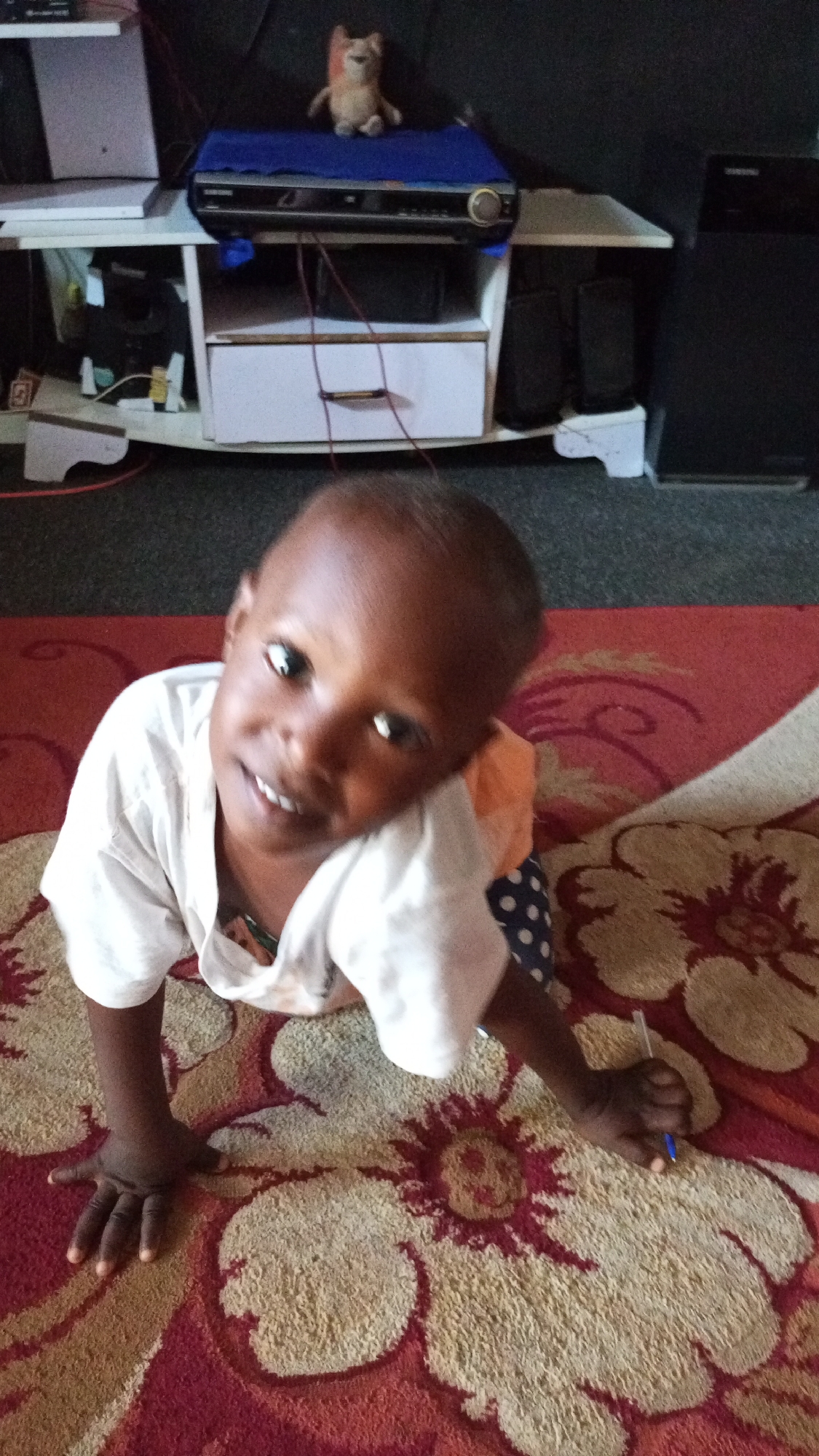 A young child with dark skin and short dark hair is the central subject of the image, looking directly at the camera with a bright smile. The child is wearing a white short-sleeved shirt and patterned bottoms (dark with white polka dots visible on the right arm). They are positioned on all fours on a patterned rug, with their left hand open on the rug and their right hand holding a blue pen.
The rug features large, abstract floral or swirl designs in shades of red and beige/cream. In the background, behind the child, is a light-colored wooden or laminate entertainment stand or small cabinet. On the top shelf of this stand, a blue fabric covers a black electronic device, which has a "SAMSUNG" logo visible on its front. A small, light-brown stuffed animal, resembling a fox or dog, sits on the blue fabric. Wires are draped over and around the stand. To the right of the stand, a large black speaker is partially visible. The background further back appears dark, suggesting an indoor setting, likely a living room or entertainment area.
The scene conveys a candid moment, with the child appearing happy and engaged, possibly during playtime.
