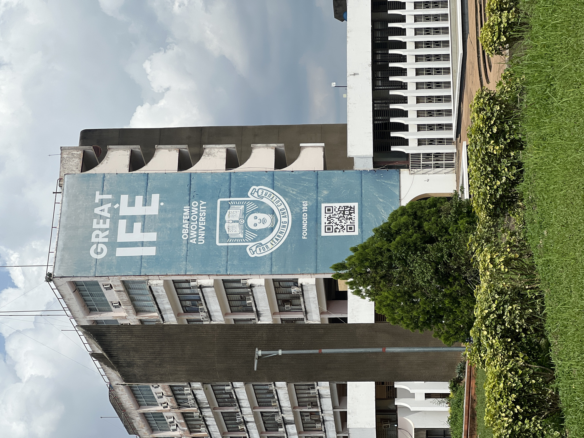 The image shows the Obafemi Awolowo University in Ife, Nigeria. A large blue banner with white text reading "GREAT IFE" is displayed on the side of a building. Below the text is the university's crest and the words "FOR LEARNING AND CULTURE" and "FOUNDED 1961". A QR code is also visible on the banner. The building is multi-story with visible windows and air conditioning units. In front of the building, there is a street light, lush green trees, and manicured bushes. The sky is partly cloudy.