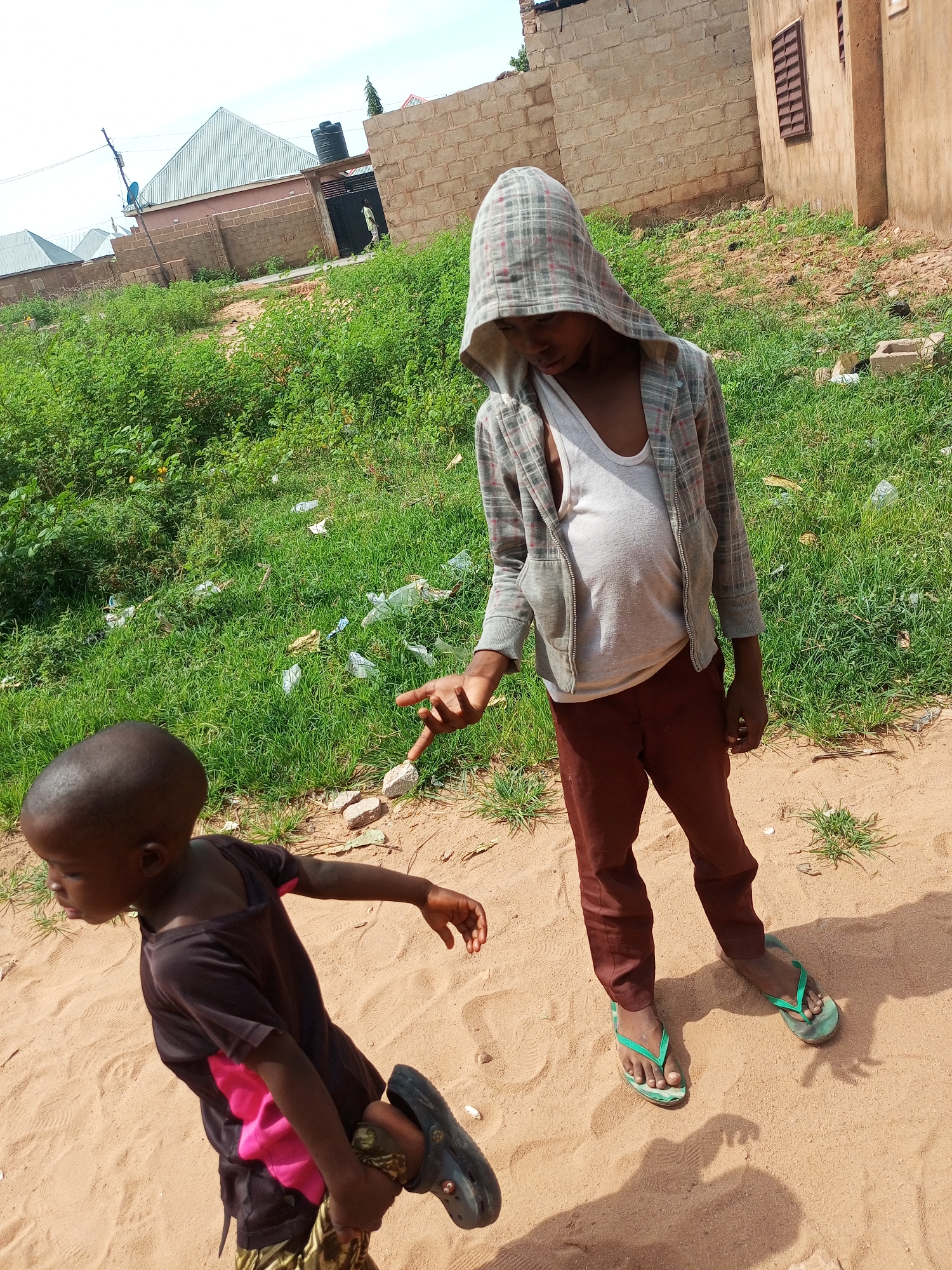 In a dusty, sandy area adjacent to a brick building with barred windows in the city of Tempure, Nigeria, two young individuals are present. The person on the right, wearing a grey plaid hooded jacket over a white tank top and brown pants, is standing and extending their right index finger towards a small, light-colored stone on the ground. Their right foot is clad in a green flip-flop, and their left foot is bare. To the left, a younger individual with a shaved head, dressed in a dark t-shirt with a pink underlayer and patterned shorts, is bending over and holding their left leg. This individual is wearing a dark sandal on their right foot. The ground is covered in sand and scattered with small stones and debris, with patches of green grass growing in the background. The environment suggests a residential or semi-developed area.