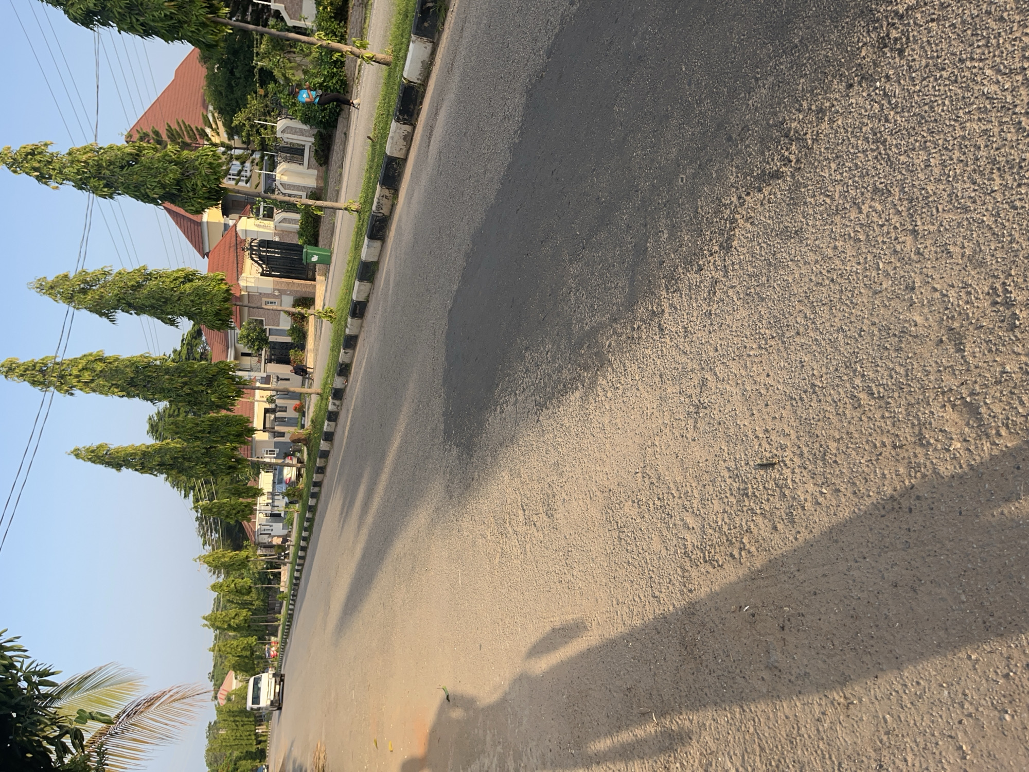 A low-angle, eye-level shot captures a residential street in Galadimawa Village, Nigeria, bathed in the warm, low light of what appears to be late afternoon or early morning. The road, paved with asphalt, dominates the foreground, its surface textured and showing signs of wear. A prominent shadow of a person, cast from the left side of the frame, stretches across the road, indicating the direction of the light source. To the right, a black and white curb borders the road, beyond which lies a well-maintained lawn and a sidewalk.
Lining the sidewalk are several tall, slender trees with lush green foliage, their branches reaching towards the clear blue sky. Behind the trees, a row of houses with red-tiled roofs and light-colored walls suggests a developed residential area. These houses feature intricate fences and gates, some with ornate metalwork, and manicured gardens. A white van is visible in the distance on the left side of the road, traveling away from the viewer. Two individuals are present; one is walking along the sidewalk to the right of the frame, and another appears to be in the distance, near one of the gates. The overall impression is one of a peaceful, affluent neighborhood in Nigeria.