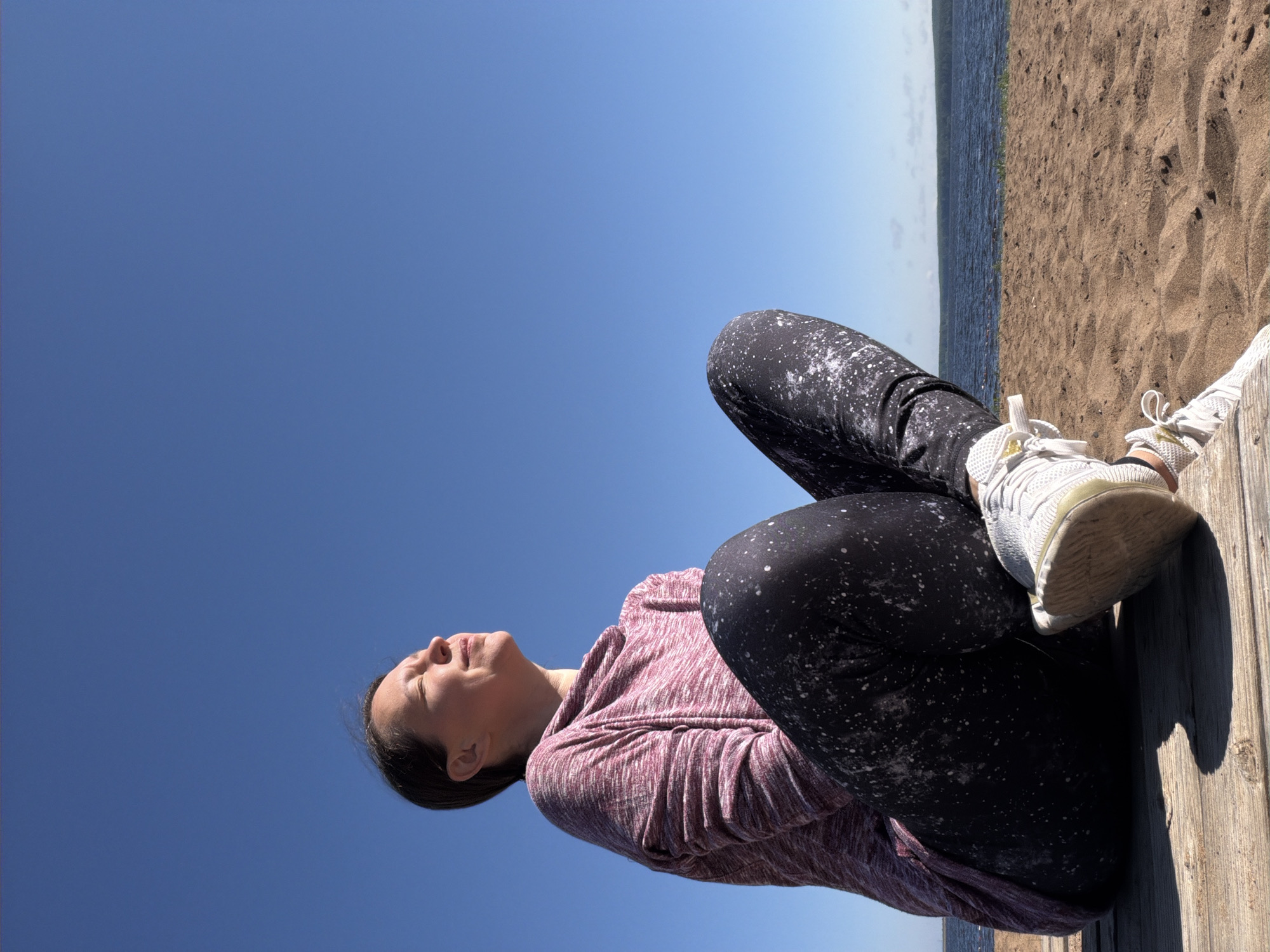 A woman sits cross-legged on a wooden deck facing the sky with her eyes closed, basking in sunlight. She is wearing a long-sleeved, marled pink and white hooded shirt and black leggings speckled with white. Her white athletic shoes are visible. The wooden deck is placed on a sandy beach, with the calm, blue water of a lake or sea in the background. A distant tree line borders the water under a clear, bright blue sky. The image appears to be taken outdoors on a sunny day. The location is not identifiable as Chaikovsky, Russia, as no specific landmarks or characteristics are present in the image to confirm this.