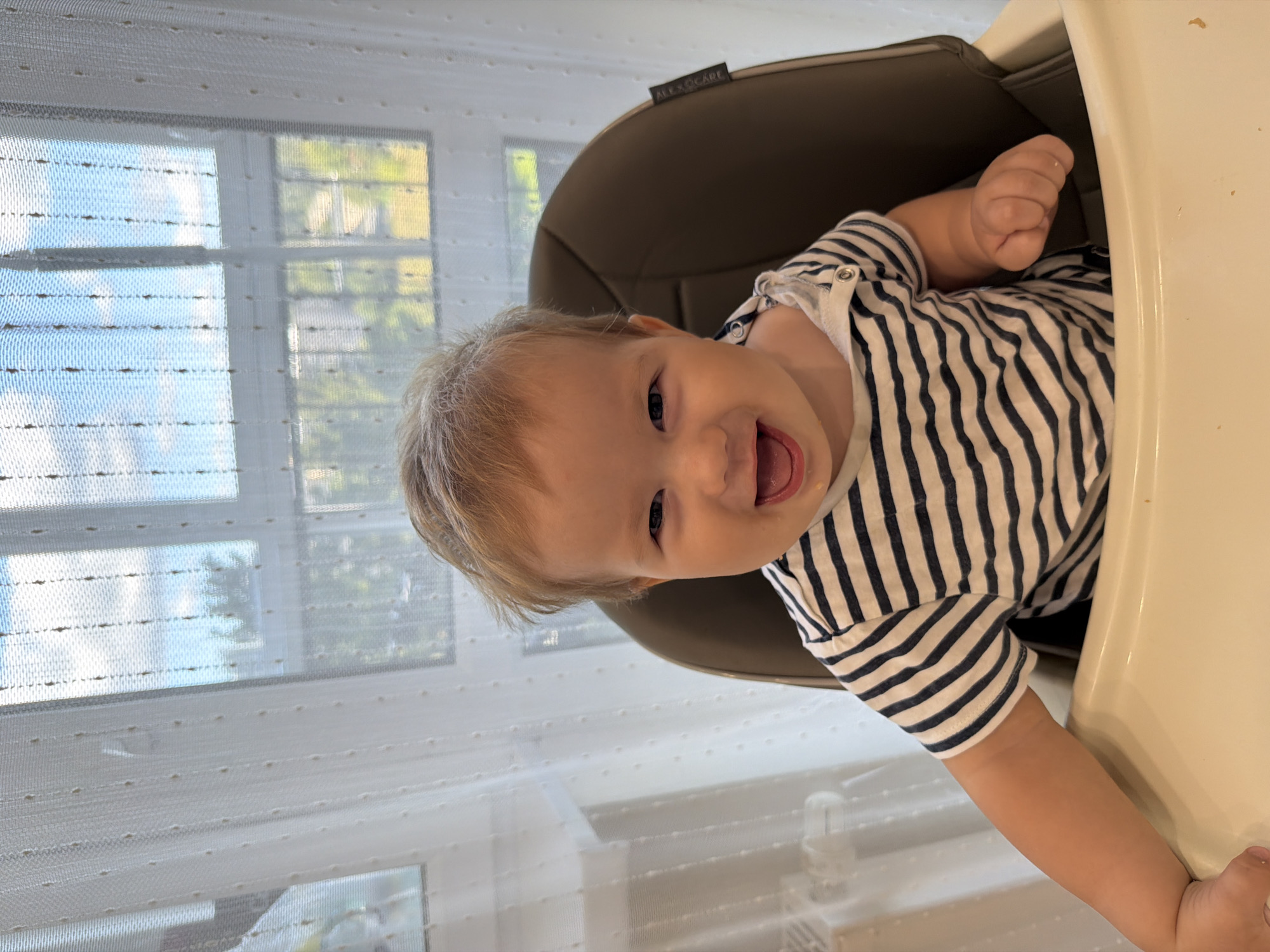 A close-up, eye-level shot captures a light-haired, fair-skinned baby smiling widely while seated in a high chair. The baby wears a navy and white striped t-shirt, and food is visible on their face and clothing. The high chair is brown and white, with a white tray. Behind the baby, sheer white curtains hang in front of a window, through which a blue sky with clouds and green trees and buildings are visible. This scene appears to be in an indoor domestic setting, likely a home, and the mention of Kazan, Russia, provides a geographical context, though no specific visual indicators of this location are present in the image itself.