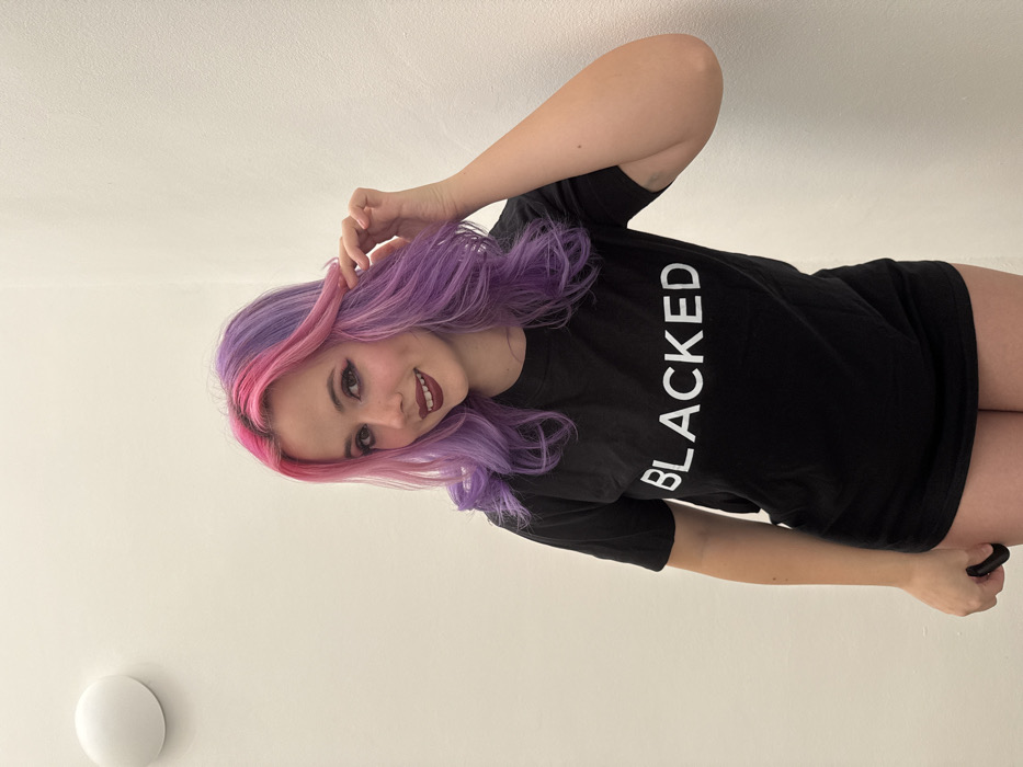 A young female, appearing to be in her late teens or early twenties, is depicted from the waist up, centered against a plain white wall. She has long hair, dyed in distinct sections of bright pink and purple, and styled with waves. Her face shows light makeup, including prominent eyeliner and dark red lipstick, and she has a slight smile while looking directly at the camera. She wears a black short-sleeved t-shirt with the word "BLACKED" printed in white uppercase letters across the chest. The t-shirt appears oversized, extending below her waist, with her lower garments not visible. Her right hand is raised, with fingers gently touching a strand of her hair near her right temple. Her left hand is lowered and holds a small, dark, unidentifiable object, mostly obscured by her thigh. A white, circular wall fixture, possibly a light or sensor, is mounted on the wall in the upper left quadrant of the image. The lighting is uniform, suggesting an indoor setting. The specific location within the city of City of Zagreb, Croatia, cannot be determined from the visual information provided.