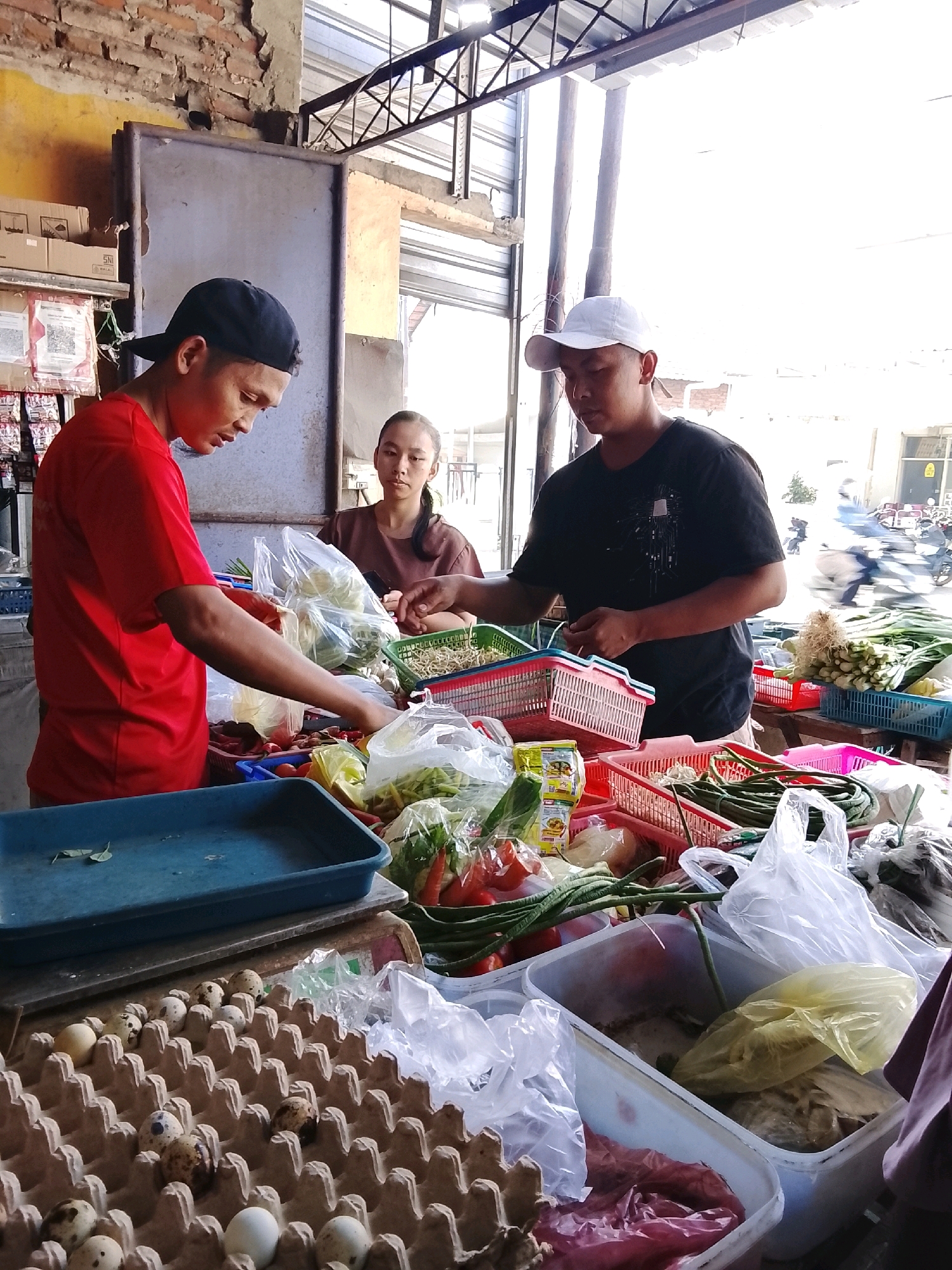 In an outdoor market stall in Dukuhtengah, Indonesia, three individuals are engaged in commerce. A young man wearing a red t-shirt and black cap, likely a vendor, is seen arranging vegetables in plastic bags. To his right, a young woman in a brown t-shirt observes the transaction. A second young man, wearing a black t-shirt and a white cap, stands behind a display of produce, appearing to be a vendor as well. The stall is filled with a variety of fresh produce, including long beans, leafy greens, tomatoes, and eggs, displayed in plastic bags and baskets. Several clear plastic containers hold additional goods. The background suggests a bustling market environment with a corrugated metal roof overhead and glimpses of other stalls and activity. The overall scene depicts a typical day at a local Indonesian market.