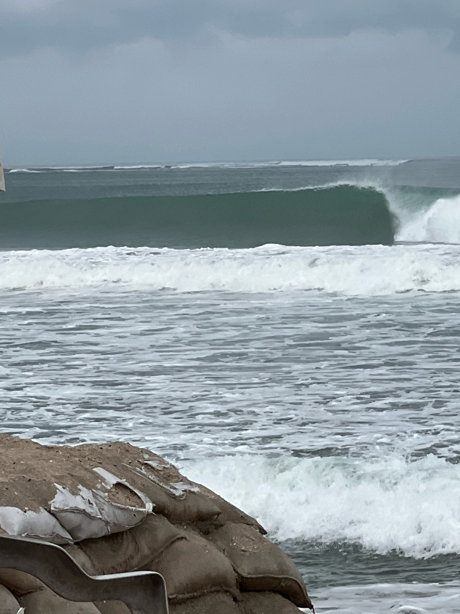 The image depicts a scene of rough seas with large waves breaking near the shore. In the foreground, a barrier of sandbags is visible, likely installed for coastal protection. The sandbags are made of burlap and some are torn, revealing white inner bags. Beyond the sandbags, the ocean is a deep teal color with white foam from the breaking waves. In the middle ground, a large, powerful wave is curling and about to break. In the background, the sea continues with more waves extending towards the horizon. The sky is overcast and a muted gray. The scene suggests a potentially stormy or high-tide condition at a beach location. This scene is consistent with the coastal erosion defense measures seen in areas like Kuta, Bali, Indonesia, particularly after periods of heavy surf or storm activity.