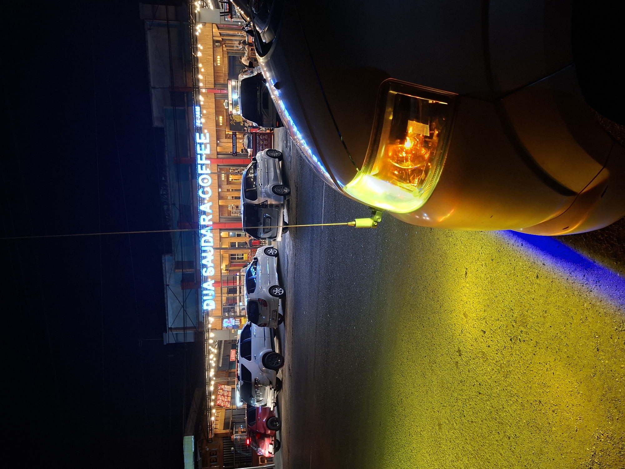 In Sinyeu, Indonesia, a night scene captures the exterior of "Dua Saudara Coffee." The foreground is dominated by the front passenger side of a light-colored vehicle, its headlight emitting a bright yellow-orange glow. The vehicle's hood curves away from the viewer, obscuring the rest of its form.
The road surface in front of the vehicle is illuminated by the car's headlights, casting a vibrant yellow and blue light pattern. Beyond the car, a row of parked vehicles, including a red hatchback, a white SUV, and two silver sedans, are visible. These cars are parked in front of a brightly lit building with a prominent neon blue sign reading "DUA SAUDARA COFFEE." The building features a warm, wood-toned facade with numerous string lights illuminating the outdoor seating area.
Several people are present, appearing to be customers and staff within the coffee shop. Some are seated at tables, while others are moving around the establishment. The overall atmosphere is one of a bustling night-time gathering at a popular local eatery. The sky above is dark, typical of nighttime conditions.