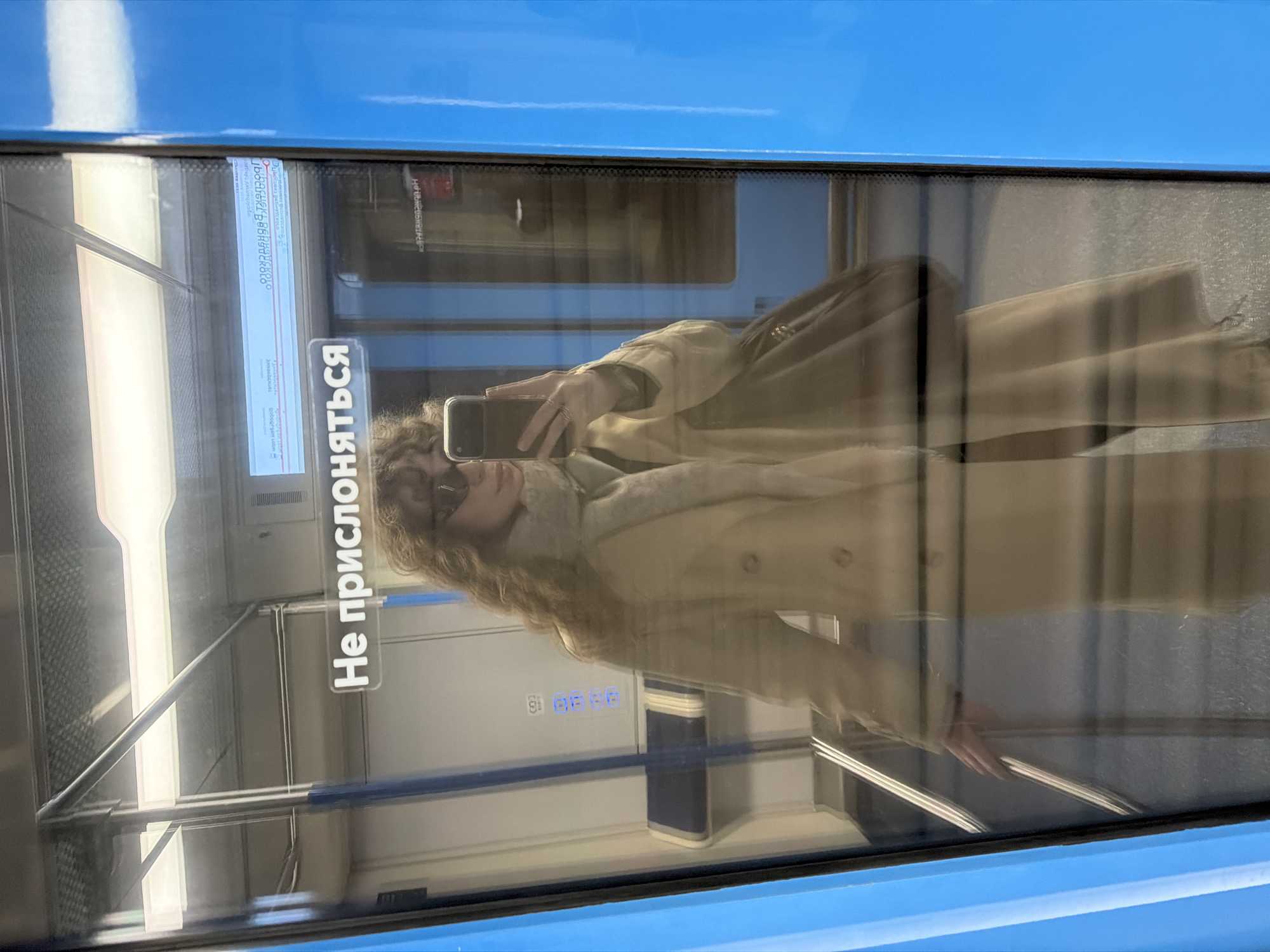 A reflection of a person in a beige coat and sunglasses taking a photo with their phone on the window of a metro train. The interior of the train is visible through the reflection, with signs written in Russian and grab rails. The image appears to be taken inside a train of the Moscow Metro.