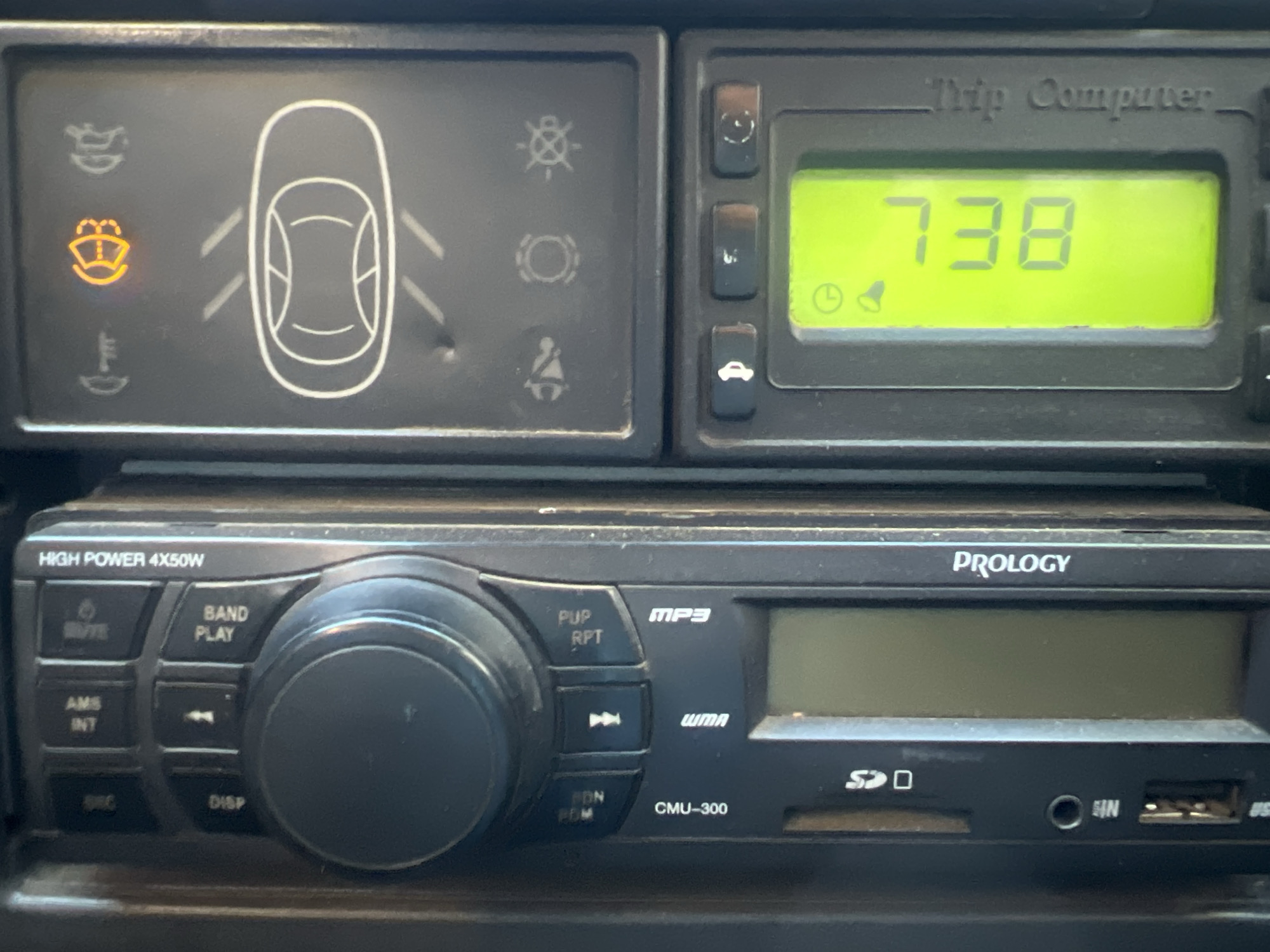 A close-up shot shows the dashboard of a vehicle. At the top left is a rectangular display with warning lights, one of which, depicting a windshield and water spray, is illuminated in yellow. To the right of it is another rectangular display, labeled "Trip Computer," showing the number "738" in green digital numerals against a yellow-green screen. Below these displays is a black car stereo labeled "PROLOGY" "HIGH POWER 4X50W" with a large central volume knob and an array of buttons, as well as slots for an SD card, auxiliary cable, and USB cable. The photograph is taken indoors, presumably inside a car. The location of Chaikovsky, Russia is unable to be verified from the image.