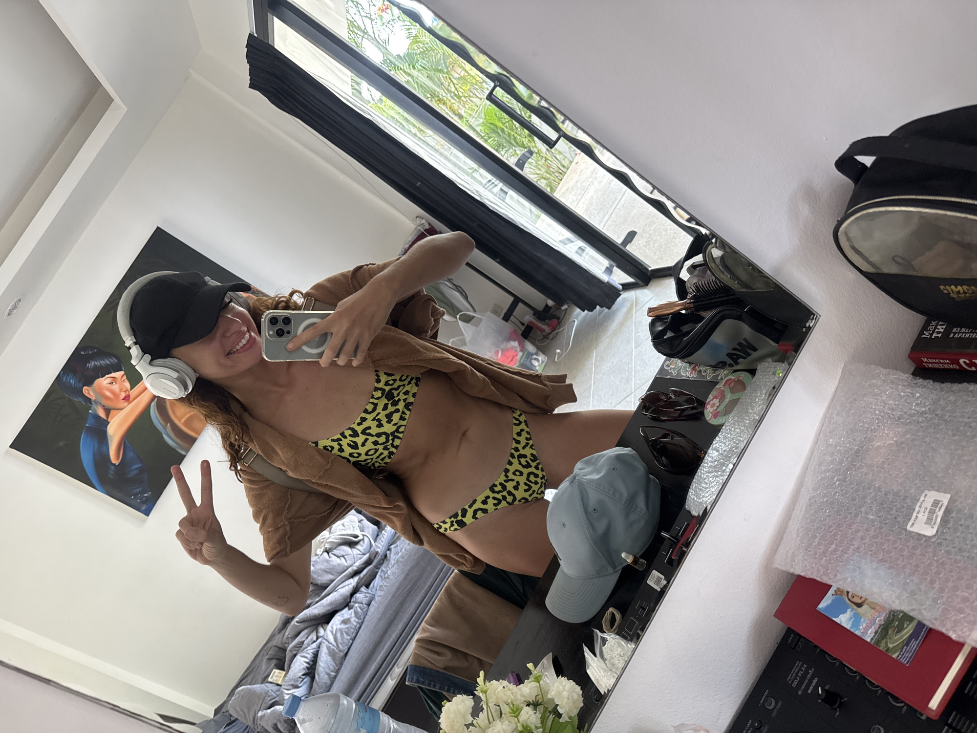 The image is a mirror selfie taken by a woman. She is wearing a black baseball cap, headphones, and a leopard print bikini with a brown cardigan. She is making a peace sign with her left hand and holding a phone in her right hand to take the picture.
In the background, a painting hangs on the wall, and a window reveals greenery outside. A bed and a water bottle are also visible in the reflection. On a surface in front of the mirror, there is a baseball cap, flowers, sunglasses, and other items. A black bag and books are to the right of the mirror.
The location is indicated by the OCR to be Baan Chaweng, Thailand, based on the brand names and text on the items shown.