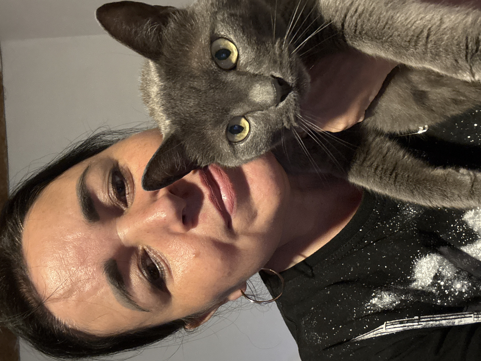 In this media file, a woman is holding a grey cat close to her face, creating an intimate selfie. The woman has dark hair and is wearing makeup, including eyeshadow and lipstick. She is also wearing a black t-shirt with a speckled white design on it. The cat has bright yellow eyes and is looking directly at the camera. The background is a plain, light-colored wall, suggesting an indoor setting. The lighting appears to be artificial, casting shadows and highlighting the subjects. The image captures a moment of affection between the woman and her pet, with both subjects displaying a relaxed and affectionate demeanor. No specific time of day or weather conditions are discernible from the image. The location is identified as Starokhalilovo, Russia.