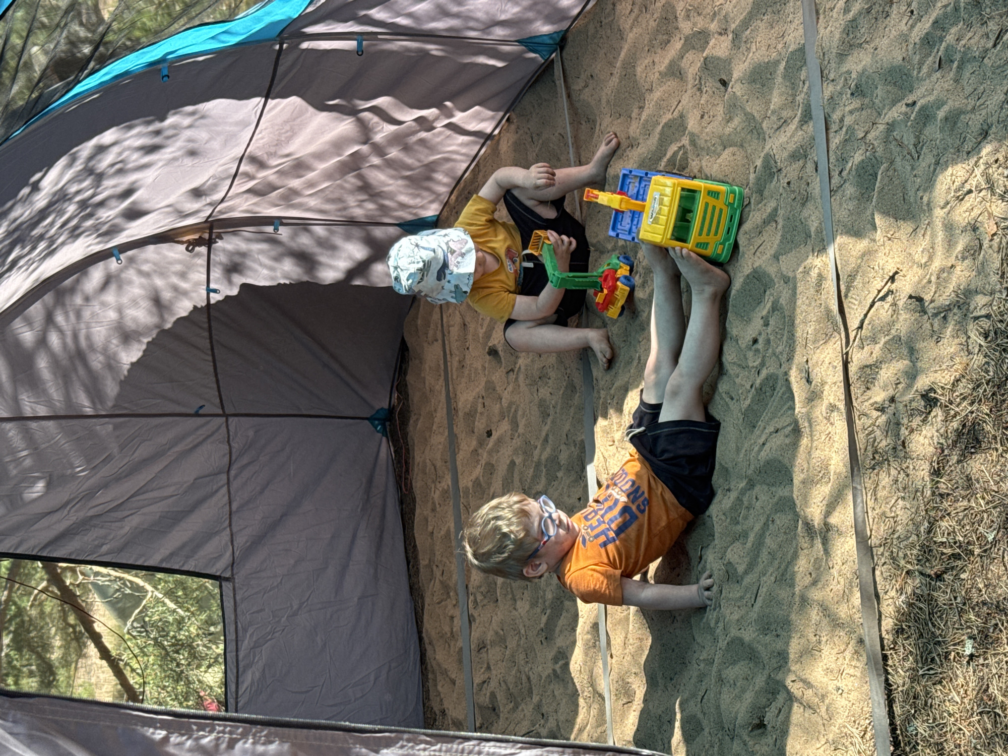 Two young children are playing in a sandpit within a tent. The child on the left, wearing an orange t-shirt and black shorts, is sitting with their legs outstretched. They have light-colored hair and are wearing glasses. The child on the right, wearing a yellow t-shirt, black shorts, and a patterned bucket hat, is kneeling and playing with two toy trucks, one green and yellow, the other yellow and blue. The sandpit appears to be an outdoor location, possibly in Ozerki (Krasnodolinskaya), Russia, with trees and foliage visible through the tent opening. The tent itself is made of grey fabric with blue accents. Sunlight casts dappled shadows on the sand and tent.