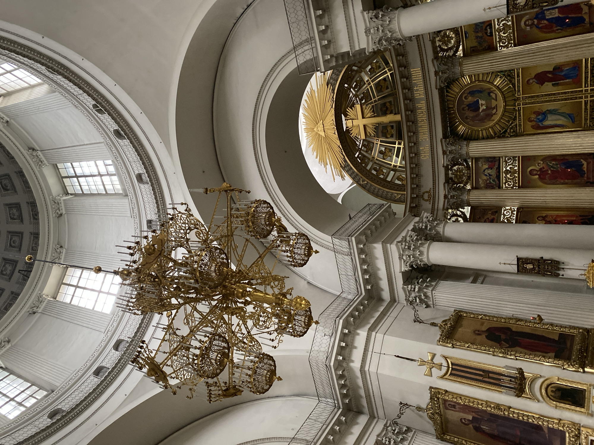 This is an interior view of Kazan Cathedral in Saint Petersburg, Russia. The image captures the grand architecture of the cathedral's dome and interior. A large, ornate golden chandelier hangs from the ceiling. The dome is supported by columns, and several windows are visible near the top, allowing natural light to enter. Below the dome, an elaborate golden structure features a cross and a sunburst symbol with an eye at its center. The walls are adorned with religious icons in ornate frames, and further down, there are depictions of religious figures and scenes. The overall impression is one of opulence and religious solemnity, typical of a large Orthodox cathedral.