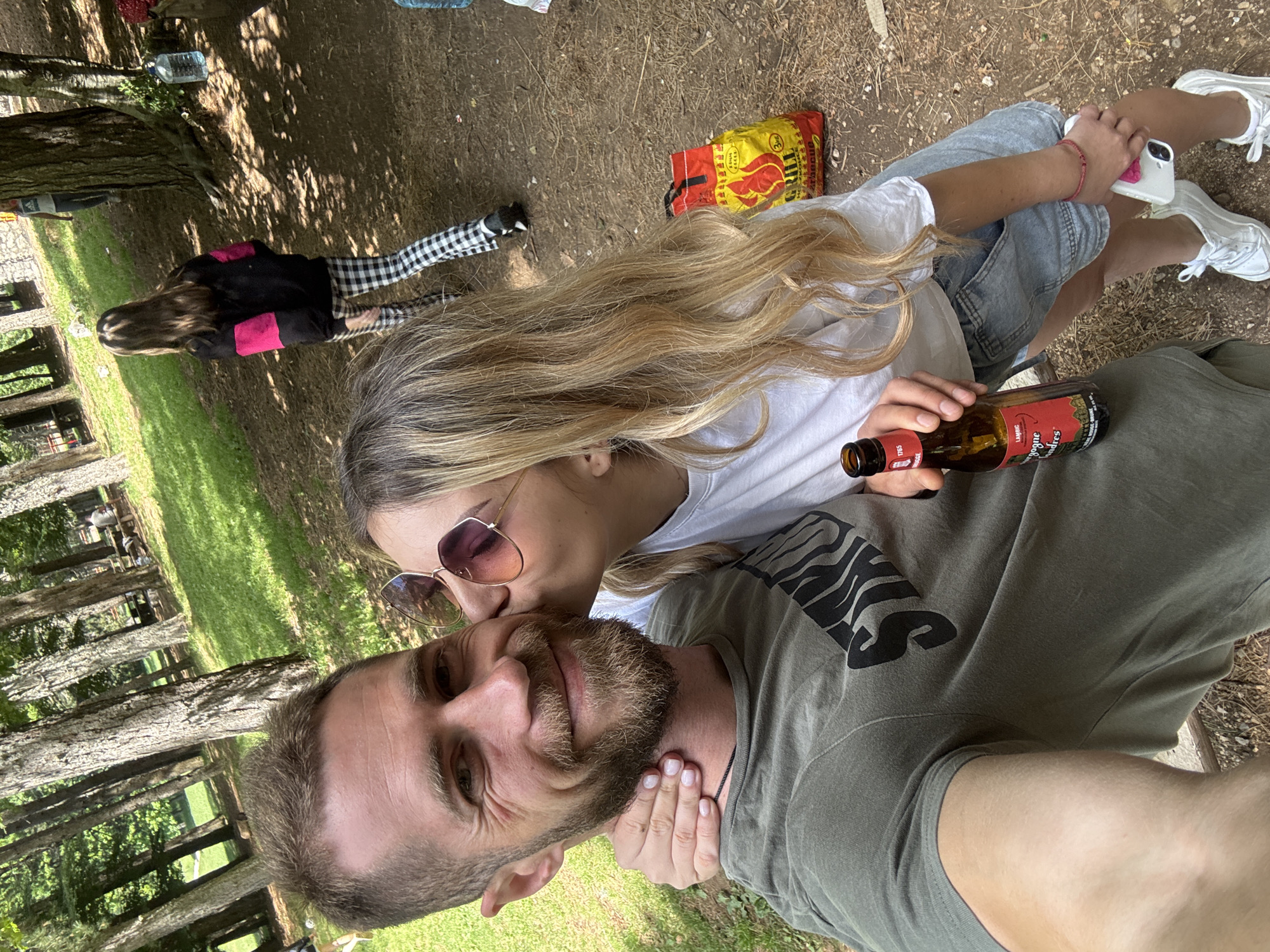The image depicts an outdoor scene in Mali Bostur, Montenegro, characterized by a wooded landscape under dappled sunlight. In the foreground, an adult male with short brown hair and a beard, wearing an olive green t-shirt, is taking a selfie and smiling. An adult female with long blonde hair, wearing sunglasses on her head, a white t-shirt, light blue denim shorts, and white sneakers, is kissing his right cheek. Her right arm is around his neck, and her left hand, adorned with a red bracelet, holds a white smartphone near her thigh. The male holds a brown glass bottle, labeled "OUDE KRIEK 1785 VANDERGHINSTE BOURGOGNE DES FLANDRES LAMBIC BRUINE," in his right hand.
In the midground, a young female with long brown hair, wearing a black long-sleeved shirt with pink shoulder panels and black and white checkered pants, walks away from the camera deeper into the wooded area. Behind her, another female in a white t-shirt and light pants is partially visible near a tree. A male figure is seated at a picnic table in the far background to the right. The ground consists of dirt, scattered leaves, and patches of green grass. Two large clear plastic water dispenser bottles and a red and yellow bag labeled "GRILL BARBECUE" are visible on the ground to the right of the central figures. A stone wall structure is partially visible through the trees in the background.