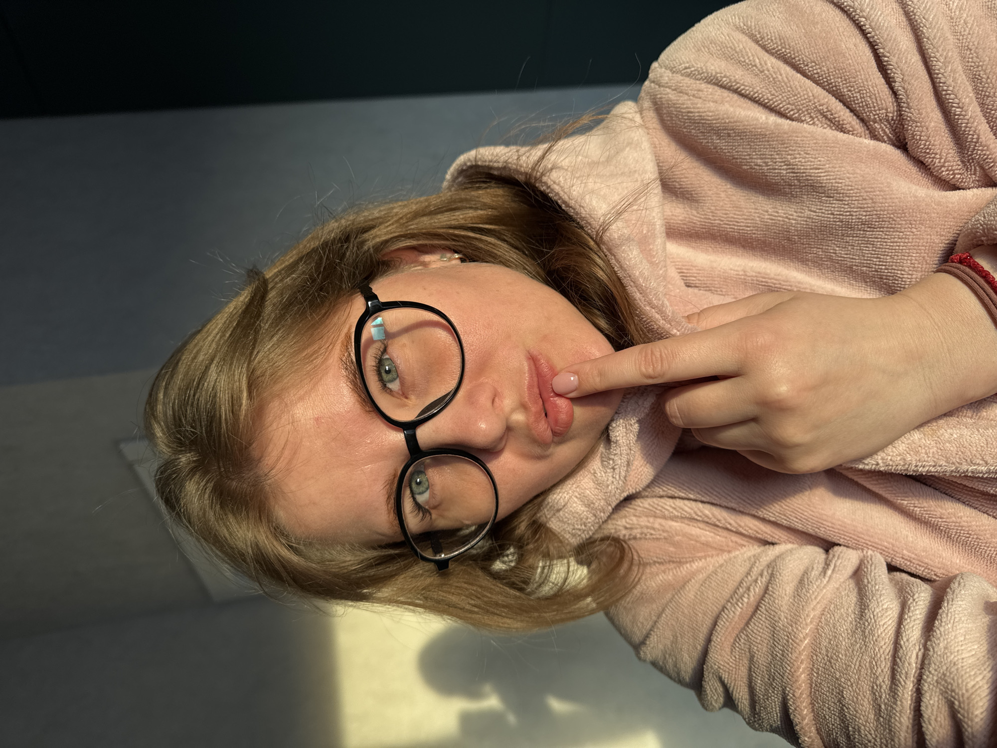A fair-skinned person with light brown hair and green eyes wearing round, black-rimmed glasses and a light pink, hooded sweatshirt is seen from a close, angled perspective. They are touching their lower lip with their index finger. The background is a neutral grey color. The location cannot be determined as being in Ферма, Russia, based on the visual information provided.