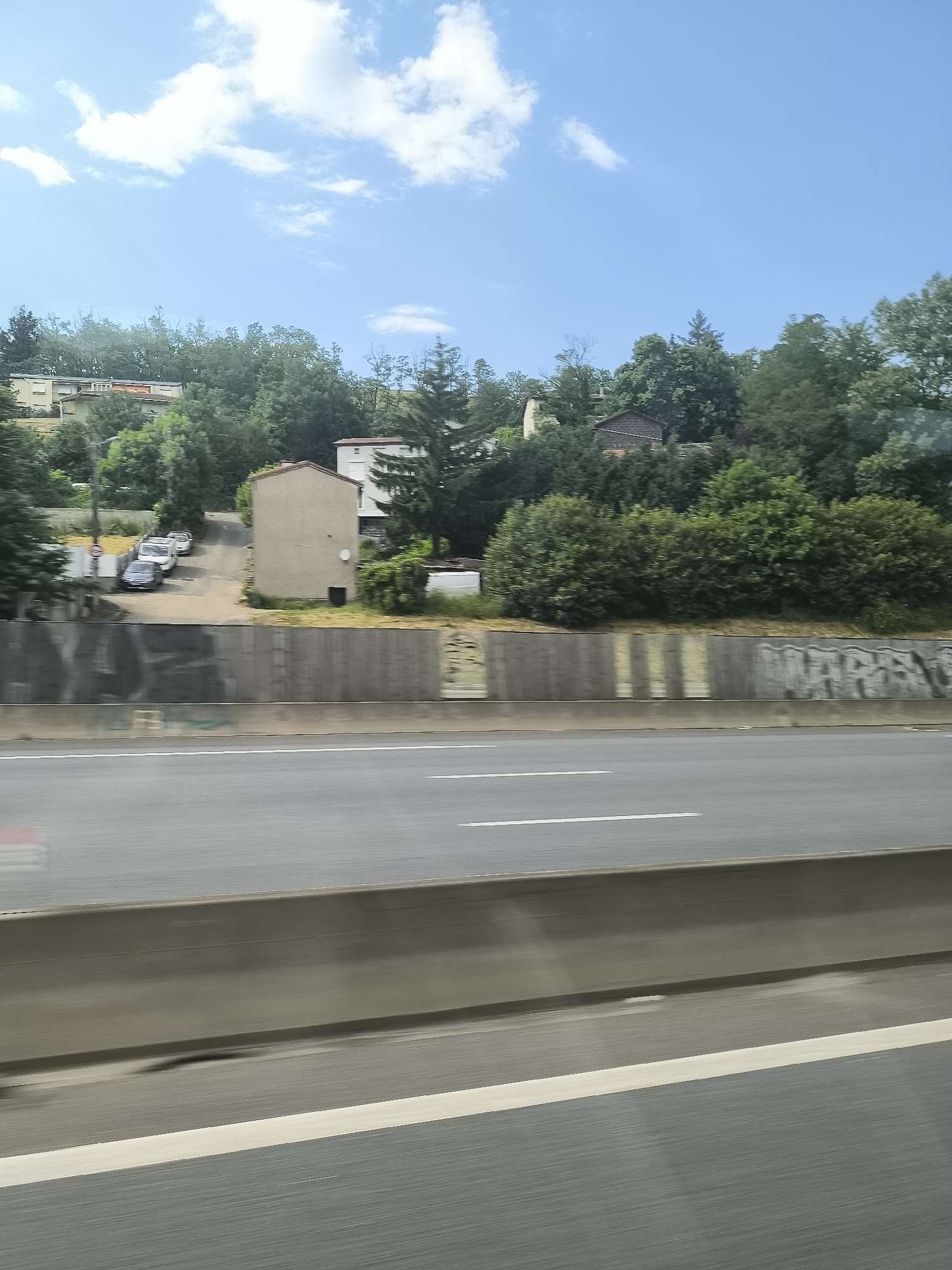 The image depicts a view from a moving vehicle, likely on a highway or motorway, in Saint-Jean-Bonnefonds, France. A concrete barrier separates the vehicle from the road. Beyond the barrier, there's a retaining wall with variations in color and graffiti markings. Above the wall, a residential area is visible, featuring several houses situated on a hillside with dense green trees and shrubbery. The sky is blue with scattered clouds. Several parked cars are seen in front of one of the houses.