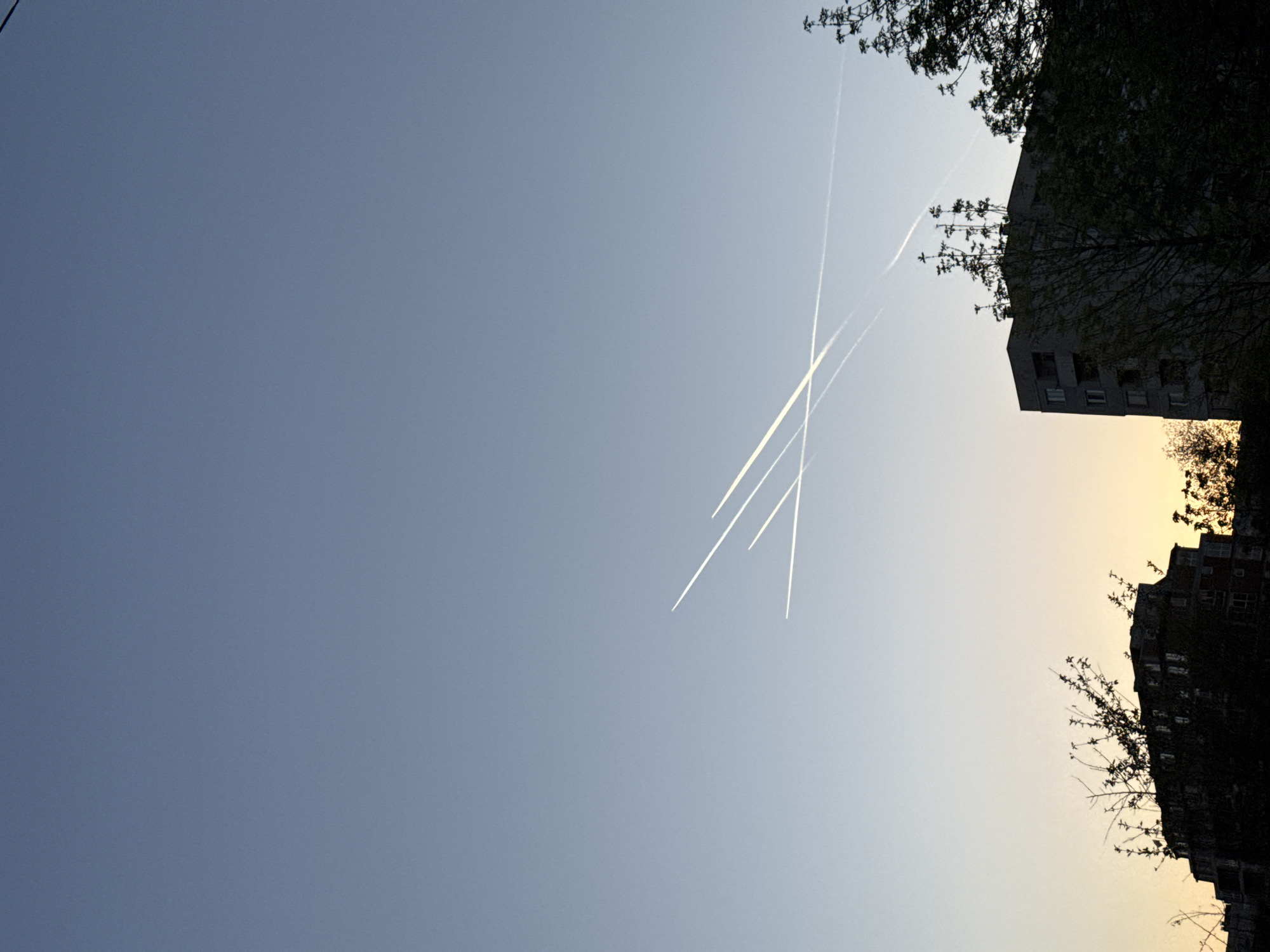 The image, taken in Sofia, Bulgaria, features a view of the sky at dusk. Several contrails from airplanes crisscross the sky, marking their paths. Buildings with trees are silhouetted against the gradient sky, where light fades from a warmer tone near the horizon to a cooler blue.