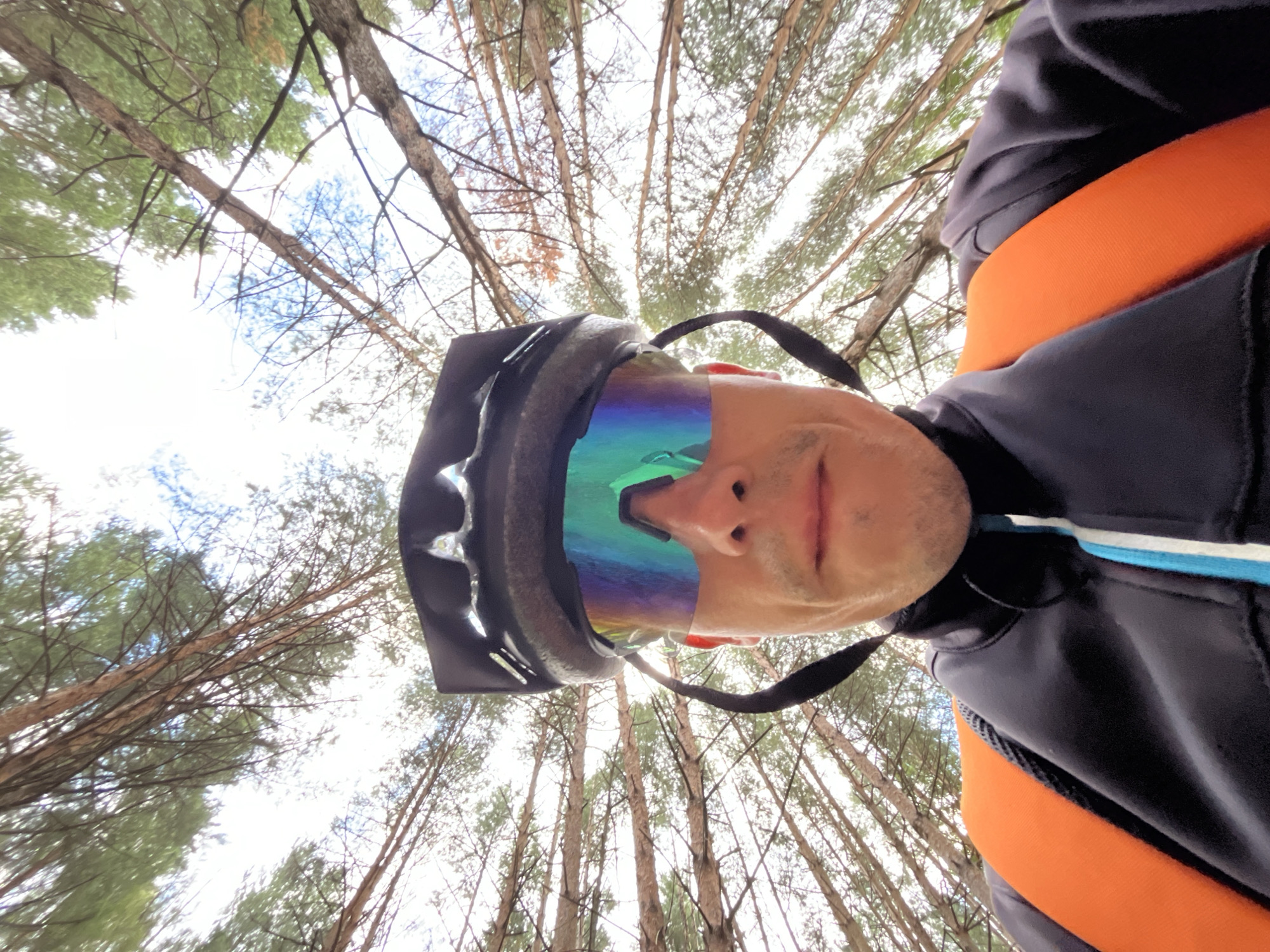 This is a close-up, low-angle, selfie-style photograph taken by a person wearing a black helmet and iridescent cycling glasses. The person is wearing a black jacket with blue and white trim and an orange backpack. The background consists of a forest with tall trees, and the sky is visible through the branches. The image was taken in the city of Chaikovsky, Russia.
