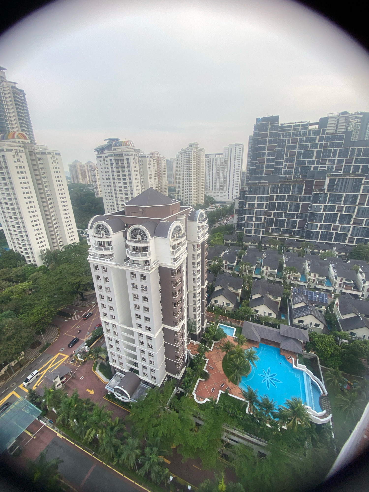 The image is a high-angle aerial view of a residential area in Kuala Lumpur, Malaysia, characterized by a mix of high-rise buildings and landed properties. In the foreground, a modern, multi-story condominium with a distinctive brown and white facade stands prominently. It features arched windows and balconies. Adjacent to this building is a large, vibrant blue swimming pool shaped like a starburst, surrounded by a red brick patio and lush green landscaping. Beyond the condominium and pool, rows of detached houses with dark tiled roofs are visible, some with swimming pools and solar panels. In the background, several other high-rise apartment buildings of varying architectural styles and heights dominate the skyline, indicative of a dense urban environment. Trees and greenery are interspersed throughout the scene, suggesting a well-developed urban landscape with pockets of nature. A street with cars can be seen at the bottom left, indicating the presence of transportation infrastructure. The sky is overcast, suggesting a cloudy day.