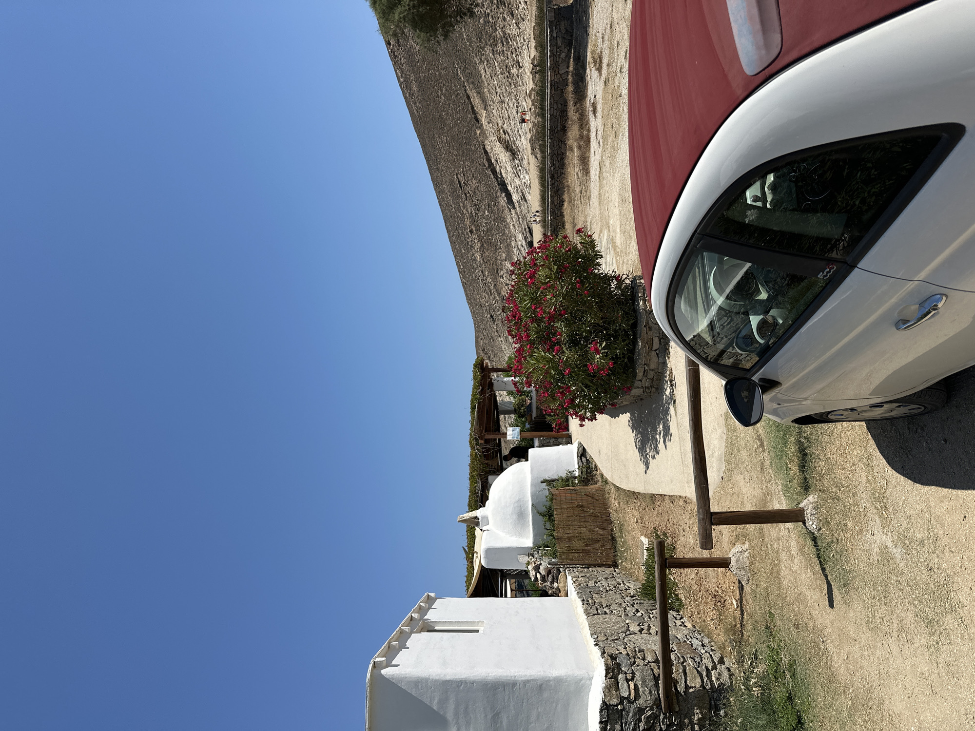 In Ano Mera, Greece, a white Fiat 500 with a maroon soft top is parked on a dirt path next to a stone wall. To the left, a white cubic building with a stone foundation and a small window is visible. Further back, a white domed structure with a small finial on top sits next to a covered seating area with a vine-covered pergola. A vibrant bush with red flowers is in bloom near the path. In the background, a sandy hillside slopes upwards under a clear blue sky. Several people are scattered on the beach in the distance. Wooden posts with a horizontal railing are positioned along the path.