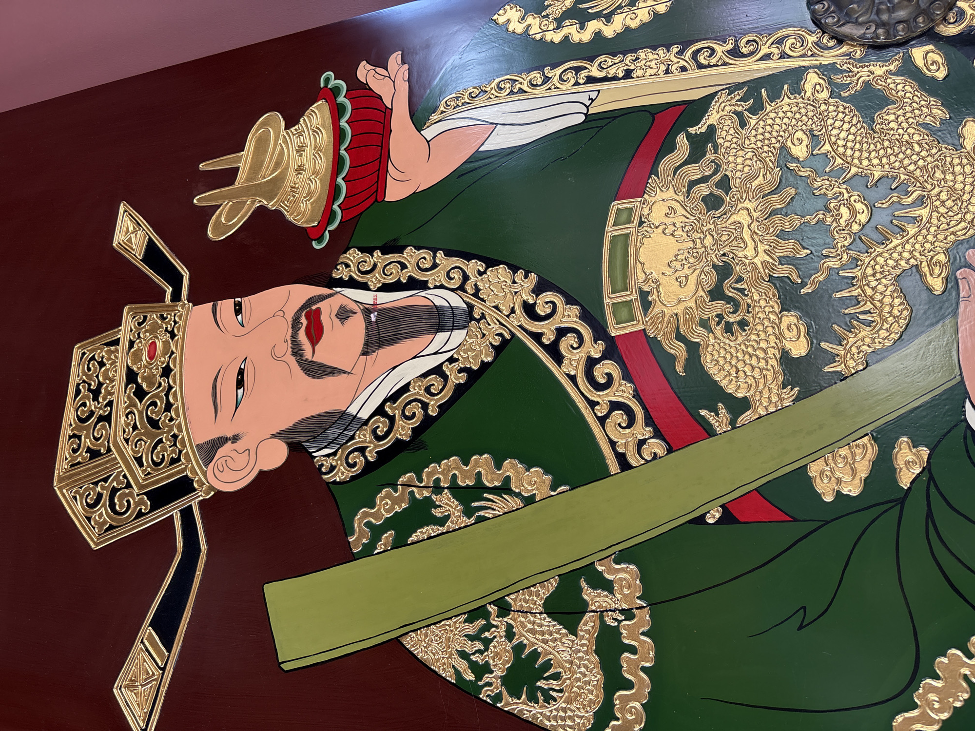 This image is a detailed painting of a man dressed in traditional East Asian attire. The man is depicted wearing an elaborate green robe with intricate gold dragon motifs and ornate gold trim around the edges. He has a dark beard and mustache, a red mouth, and his eyes are narrowed, looking forward. He wears a tall, black hat adorned with gold detailing and a small red gem. In his right hand, he holds a golden, goblet-like object with a rabbit-like figure on top. In his left arm, he holds a long, green, rectangular object. The background is a deep red-brown.
This painting is located at the Shanghai Museum in Shanghai, China.