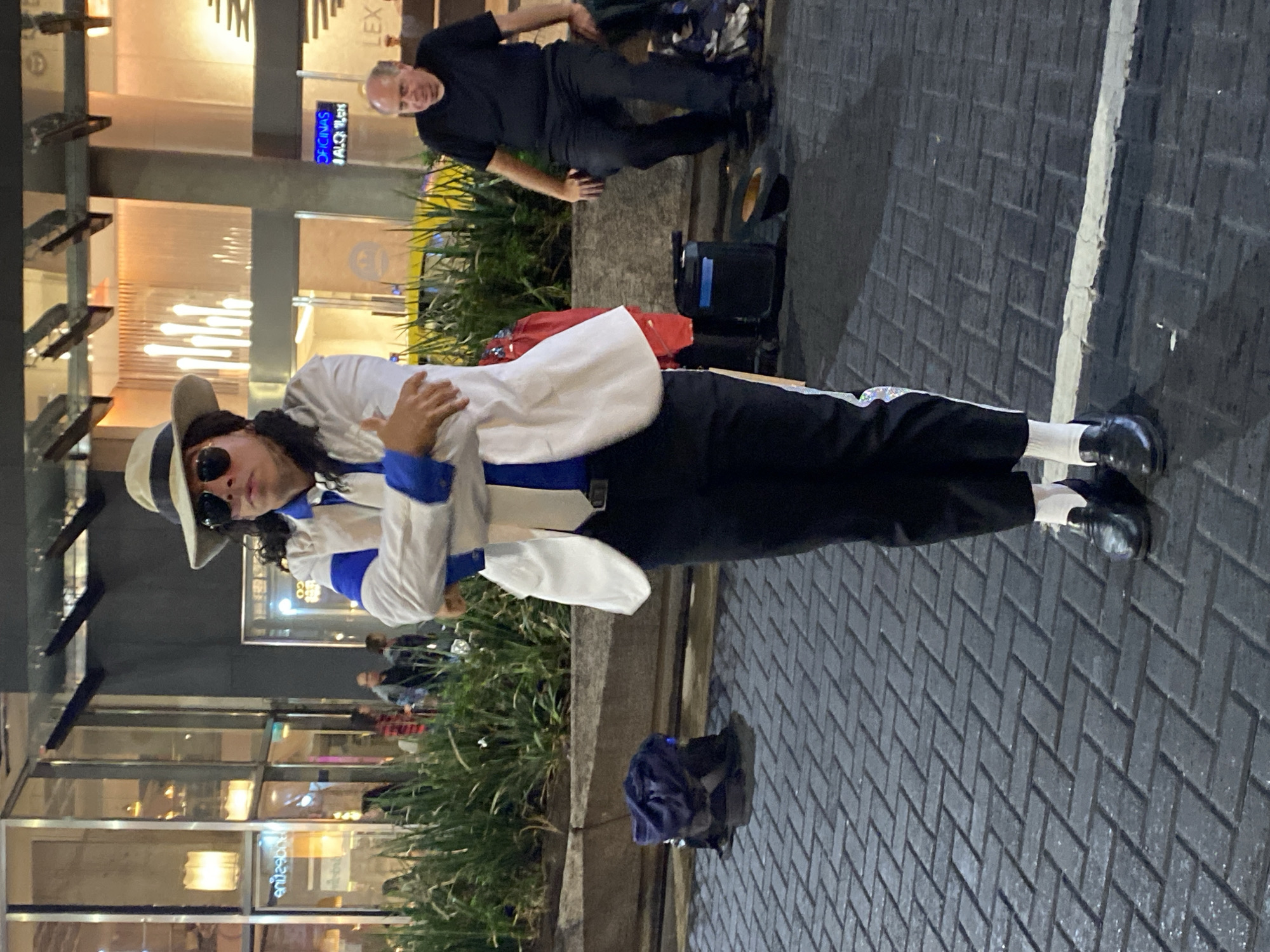 This image depicts an urban outdoor scene in the Microcentro (financial district) of Buenos Aires, Argentina, observed at night or dusk under artificial lighting.
A person dressed as Michael Jackson stands centered on a dark grey paved walkway composed of interlocking rectangular pavers. This individual wears a light-colored fedora, dark sunglasses, a white blazer over a blue shirt, a white tie, black trousers with a prominent white vertical stripe on the outer seam, white socks, and black loafers. Their arms are crossed over their chest, and they maintain a forward gaze.
To the right of the performer, a man in a black t-shirt and dark trousers is seated on a concrete planter or ledge. Beside him, on the paved ground, are a black portable speaker/amplifier and a dark fedora hat, positioned upright. A dark backpack, with a blue item resting on top, is visible behind the performer, near the planter. A red garment is also partially visible draped near the speaker.
The background features a modern building with extensive glass facades, reflecting interior lights and architectural details. Mirrored text "enilzeboR" (Rosbez) is visible on one window, while other signs clearly display "OFICINAS ALQUILER" (Offices for Rent) and partially "LEX", suggesting commercial spaces. Green foliage, likely shrubs or grasses, grows in the planter separating the walkway from the building. A faded white line runs across the paved ground in the foreground, parallel to the viewer.