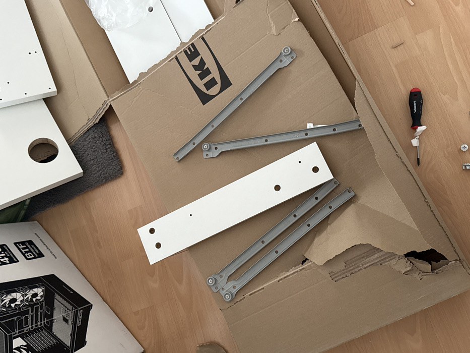 The scene is an indoor location in Vellmar, Germany, depicting a collection of unassembled furniture components and computer hardware packaging laid out on a light brown wooden floor. In the center, a large, open, brown cardboard box, featuring the "IKEA" logo on its inner flap, is visible. This box appears partially torn on its left side. Within and around the box are several white, flat-pack furniture panels of various sizes, drilled with multiple holes for assembly. Prominently featured on the cardboard box is a long, narrow white panel with multiple circular and smaller drilled holes. Adjacent to this, multiple grey metal drawer slides, each with small wheels at one end and screw holes along their length, are distributed.
In the upper left portion of the image, a white and black cardboard box for a "Sharkoon TG4M RGB" computer case is present, displaying an illustrative rendering of the product and text including "41.9 cm" and "BTF." A dark grey, shaggy rug is partially visible beneath some of the white panels and the IKEA box in the upper central area. On the wooden floor in the lower right, a screwdriver with a black and red handle, marked "WIESMANN" and adorned with a white plastic tie, lies next to several small wooden dowels and various metal fasteners, including screws and cam locks. No individuals are present in the scene.