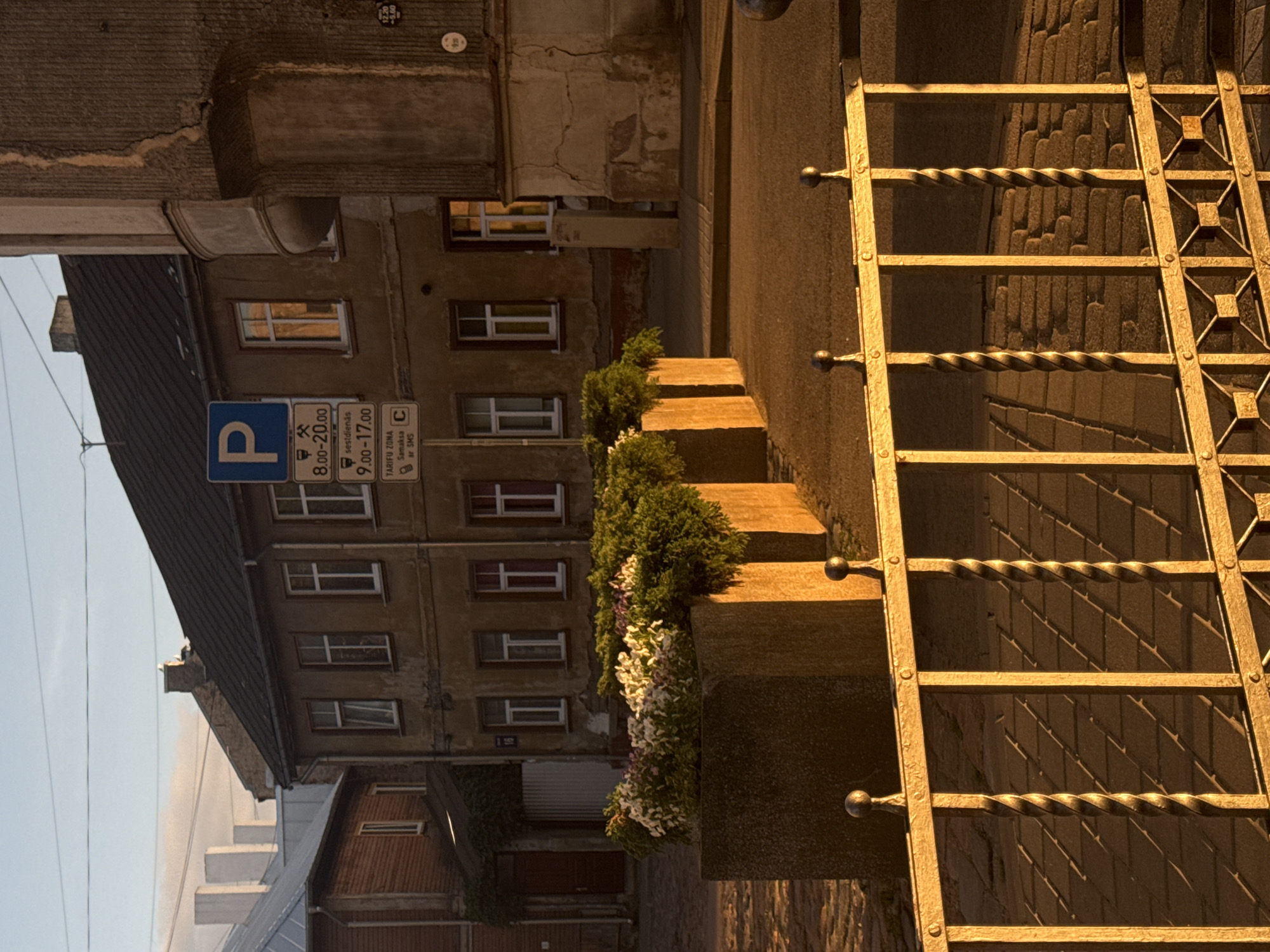 The image depicts a street scene in Riga, Latvia, bathed in the warm, low light of either sunrise or sunset. In the foreground, a golden metal fence with decorative spiral elements runs horizontally across the frame. Behind the fence, a cobblestone sidewalk is visible, casting long shadows from the fence's structure. Further back, a row of large concrete planters contains lush green plants and white and pink flowers.
The background features an old, multi-story building with a dark roof and beige walls. Windows of various sizes are visible, some with white frames and others with darker frames. One window reflects the warm light. A blue parking sign is mounted on the building, accompanied by smaller signs indicating parking hours and zones. To the left of this building, another older building with a red wooden facade and a dark roof is partially visible, with a red door and a green awning. In the far distance, the hazy outlines of modern buildings, possibly skyscrapers, can be seen against the sky, suggesting a contrast between old and new architecture within the city. The sky above is a pale blue, with faint wisps of clouds. The overall atmosphere is serene and quiet, typical of early morning or late evening in a European city.