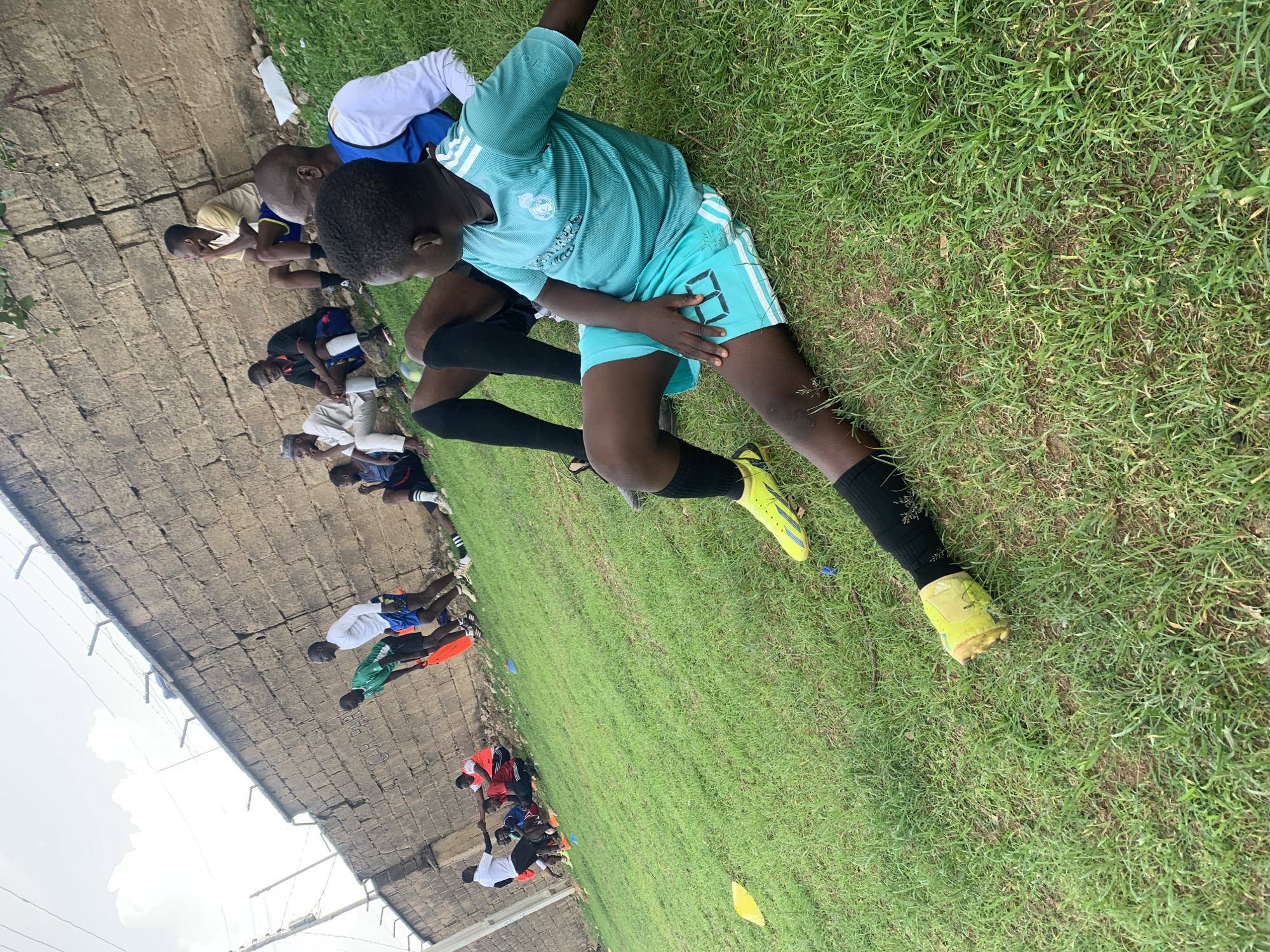 A ground-level shot depicting a row of young African men stretching on a grassy field. They are near a wall; the perspective suggests they are participating in a soccer training session. The location is outdoors in Jos, Nigeria.
The men are lined up along the edge of the field, close to a tall, weathered stone wall. They are uniformly dressed in soccer attire, including jerseys, shorts, and athletic shoes. Their jerseys are various colors, including blue, green, white, red, and yellow. Some are wearing athletic socks.
The environment includes a green grass field, and the grey, stony wall. The sky is partly cloudy.