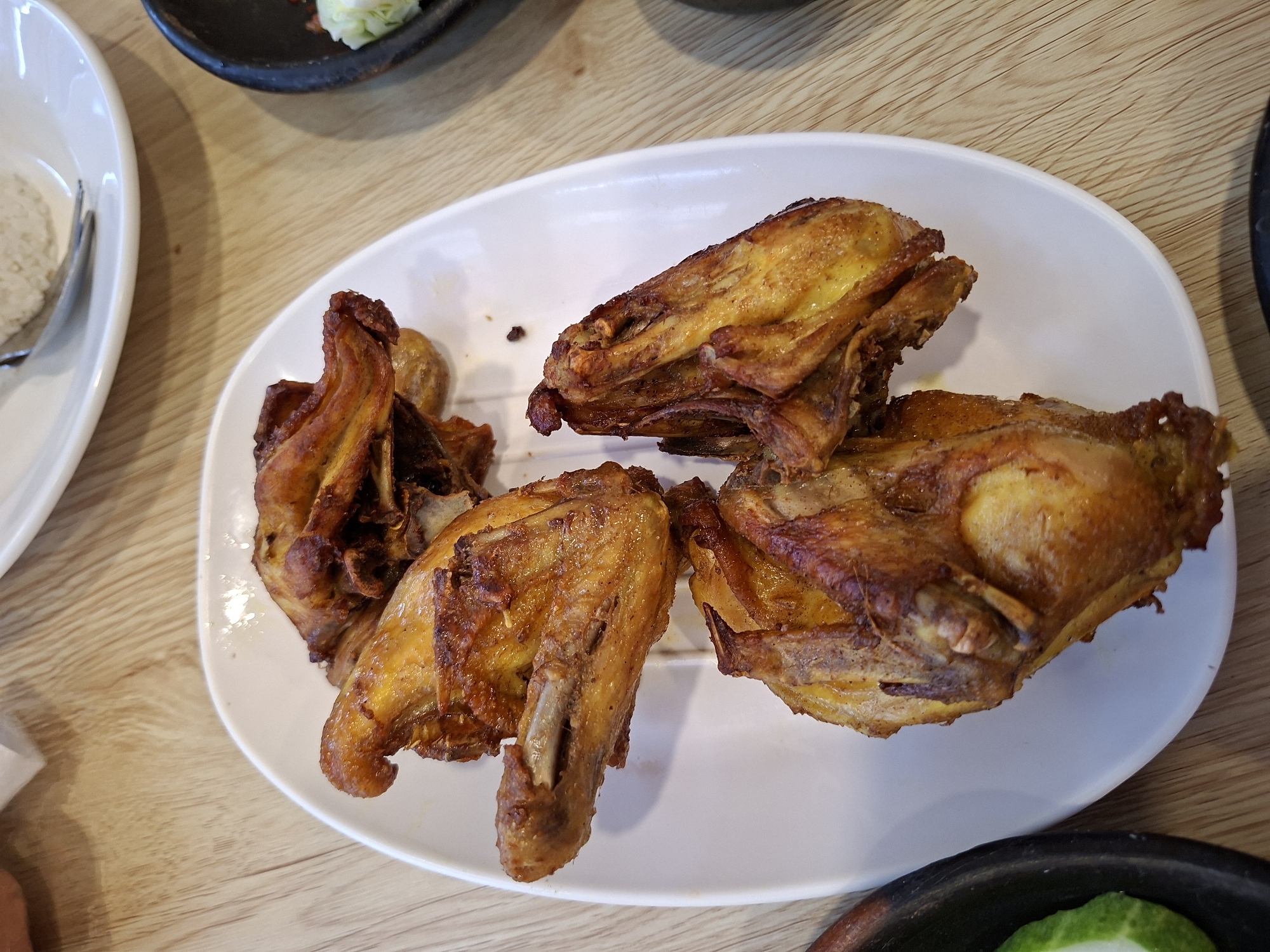 A plate of fried chicken is displayed on a wooden table in Banjaratma, Indonesia. The fried chicken pieces, appearing to be wings and a thigh, are golden brown and glistening. They are arranged on a white oval plate. In the background, a white plate with rice and a fork is visible, along with a dark bowl containing shredded vegetables. The lighting suggests an indoor setting, possibly a restaurant or home dining area.