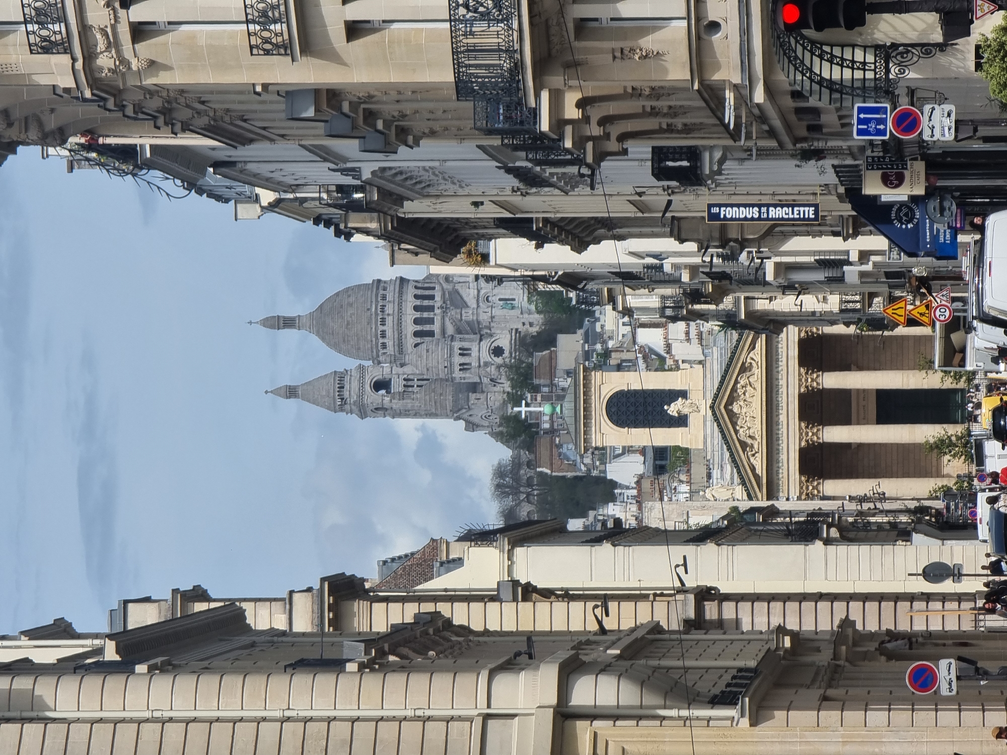 The image displays a bustling street scene in Paris, France, with the iconic Sacré-Cœur Basilica prominently in the background. The perspective looks up a narrow street, framed by classic multi-story Parisian buildings constructed from light-colored stone, featuring ornate wrought-iron balconies and traditional pitched rooftops with dormer windows.
In the middle ground, the majestic white domes and bell tower of the Sacré-Cœur Basilica rise above the surrounding urban landscape, bathed in bright daylight under a partly cloudy sky. Below the basilica, other buildings with distinctive architecture, including what appears to be a structure with classical columns and sculptural details, are visible.
The foreground is dominated by the detailed facades of the buildings lining the street. On the right, a red traffic light is illuminated, alongside various street signs. These include a blue square sign with a white arrow (likely indicating a one-way street or direction), a red circular "no entry" or "no parking" sign, and a yellow triangular warning sign above a speed limit sign displaying "30" in a red circle.
Several commercial signs are visible, clearly reading "LES FONDUS DE RACLETTE," indicating a restaurant. Other partially visible signs include "SANDWICHS CAFÉS," "COMPTOIR," and "A EMPO" (presumably "à emporter"). A sign that appears to say "G&Coiffeur" suggests a hairdresser. A white delivery vehicle or van is partially seen on the far right. The scene depicts a typical bright daytime ambiance in the city.