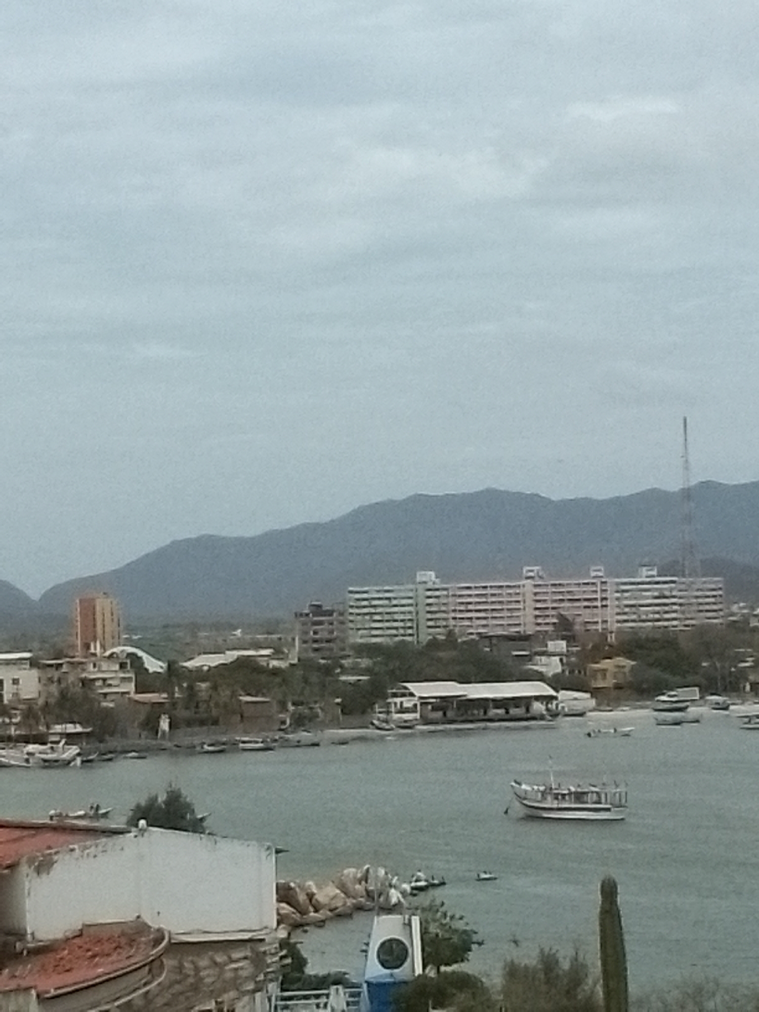 The image depicts a coastal view in Juan Griego, Venezuela, under an overcast sky. The foreground features the top of a building with a red-tiled roof and a white wall, partially obscuring the view. A bay stretches into the middle ground, dotted with several small boats and a larger tour boat in the center. On the shoreline, various low buildings and houses are visible. Behind them, a large, multi-story building complex dominates the skyline, and a tall communications tower stands to its right. In the background, mountains rise, completing the landscape. A tall cactus is visible.