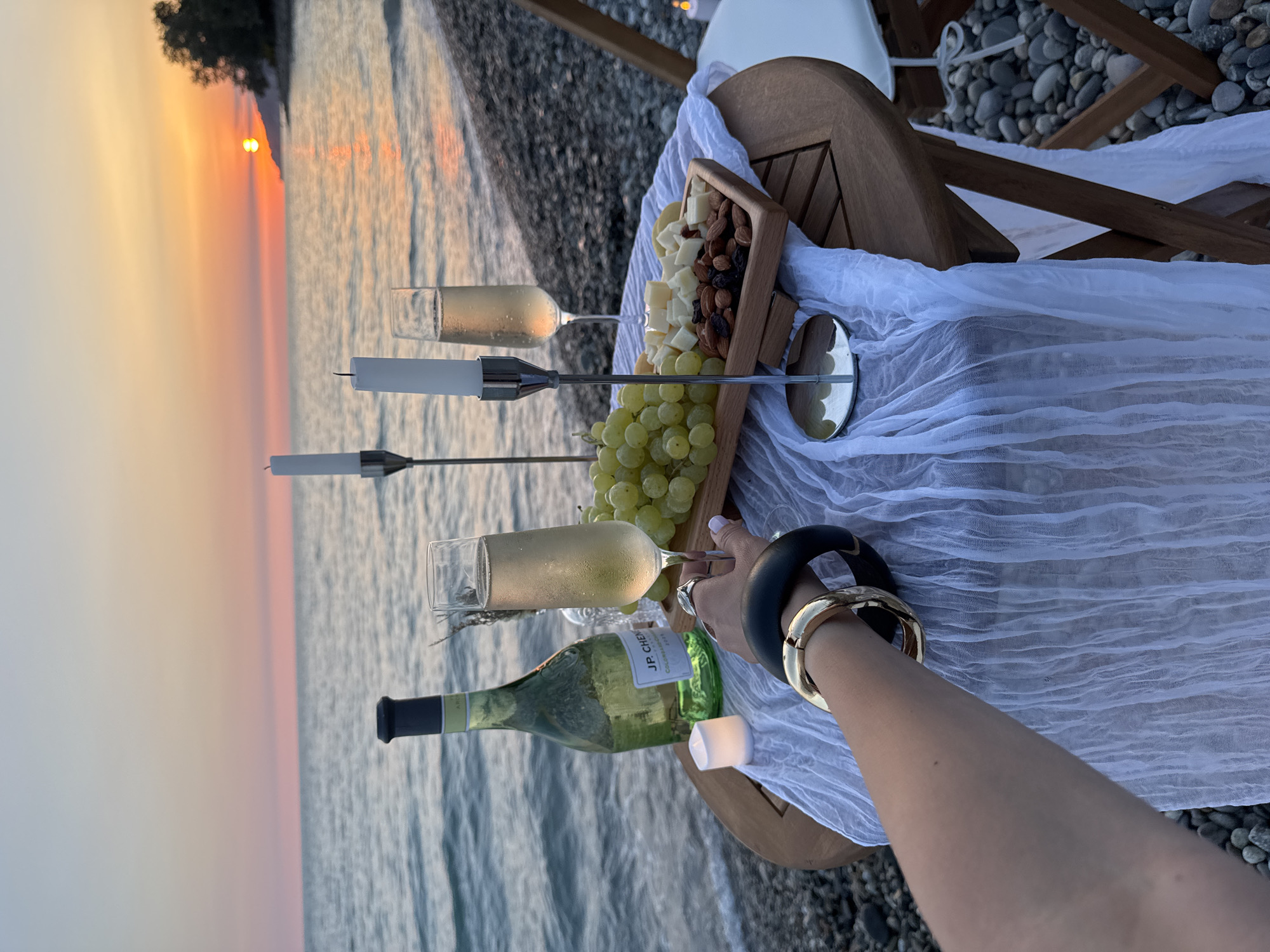 A close-up shot captures a romantic sunset picnic on the pebbled shore of the city of Sochi, Russia. A small wooden table is set with a bottle of white wine, two flutes of champagne, a bouquet of green grapes, and a wooden tray filled with cheese cubes, almonds, and raisins. Two tall white candles in silver holders stand prominently on the table. A white translucent cloth is draped over the table. In the foreground, a person's arm, adorned with gold and black bracelets, reaches towards a champagne flute. The calm sea stretches out to the horizon, where the sun is setting in a blaze of orange and pink hues. The overall atmosphere is serene and intimate.