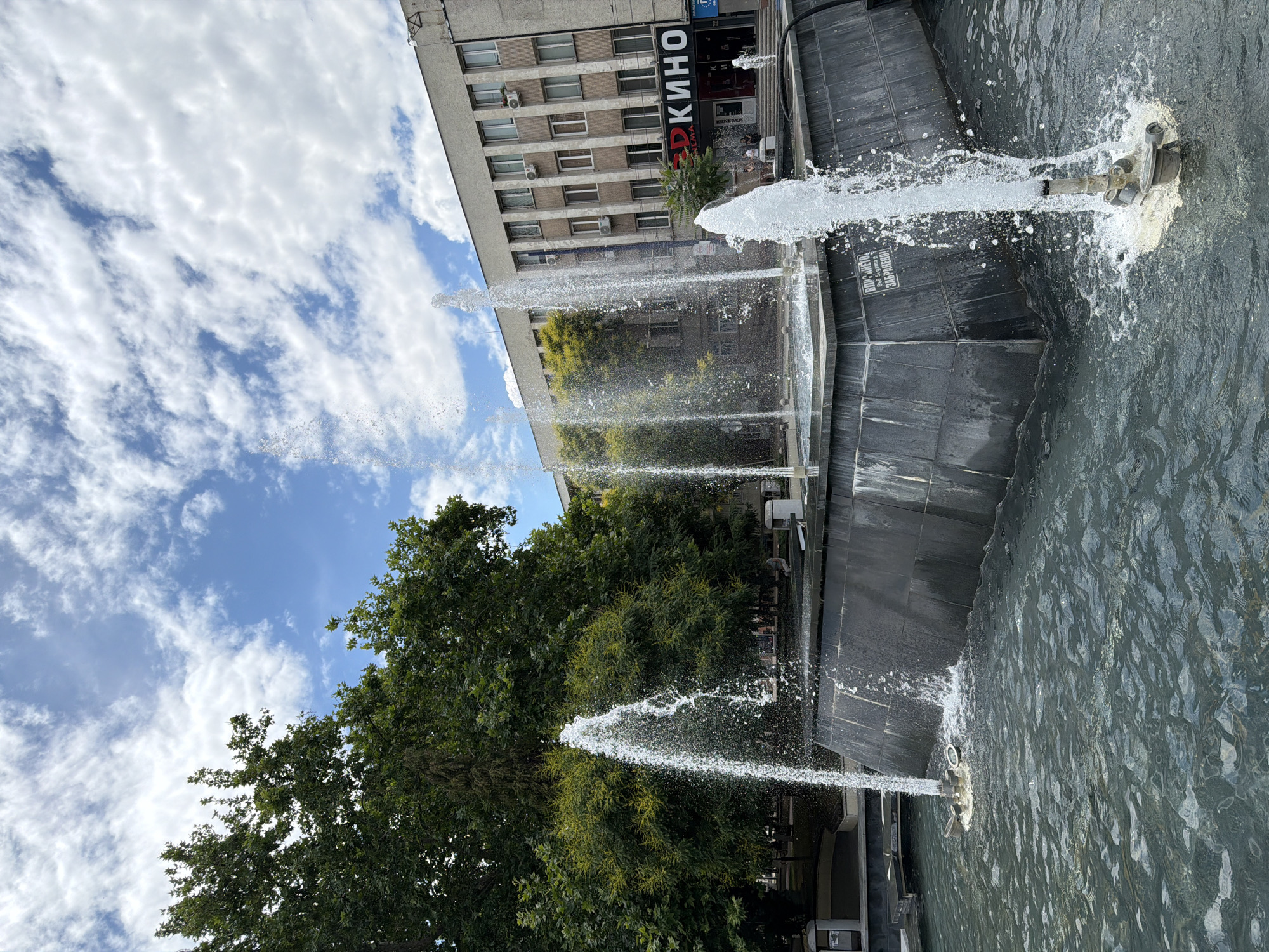 This is a wide shot of a modern fountain in an urban setting, likely in the city of Pazardzhik, Bulgaria. The fountain has a dark, multi-tiered base from which multiple water jets spray upwards. The water is clear and illuminated by the sunlight, creating a dynamic and lively atmosphere. In the background, a building with a sign that reads "КИНО" (meaning "Cinema" in Bulgarian) is visible. The building is several stories tall with numerous windows and air conditioning units. Lush green trees, characteristic of a temperate climate, flank the left side of the fountain and the building. The sky above is a mix of blue with scattered white clouds, indicating fair weather. A few individuals are faintly visible near the entrance of the cinema. The overall scene suggests a public space designed for recreation and gathering.