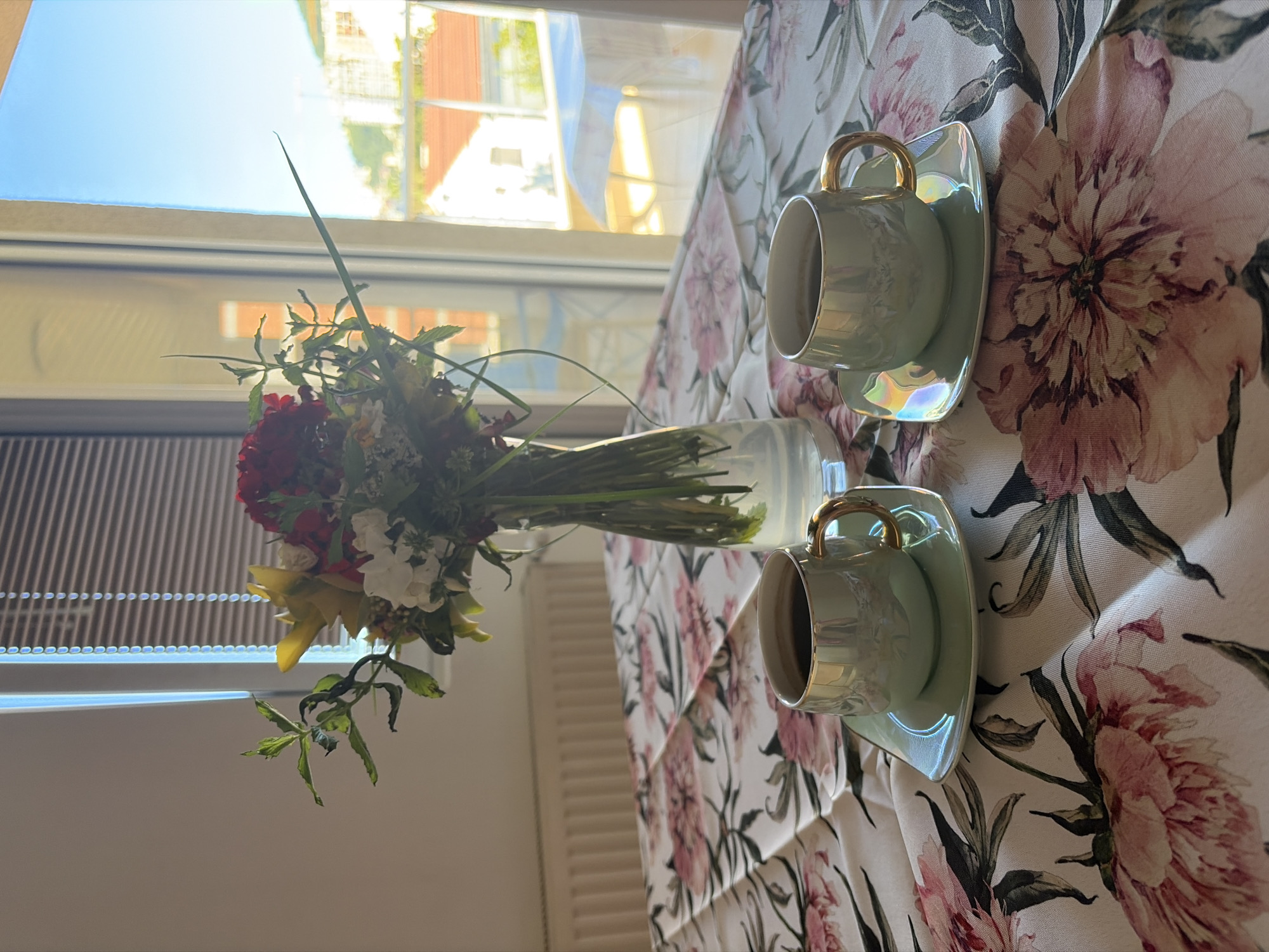 The image shows a table setting with a floral tablecloth, two light green tea cups with saucers, and a glass vase containing a bouquet of flowers. The setting is likely indoors, near a window. The window shows a glimpse of buildings and bright sky. Based on the visual information available, I cannot conclusively determine the location to be Budva, Montenegro. More specific visual cues unique to the city's architecture or landscape would be needed for that level of detail.