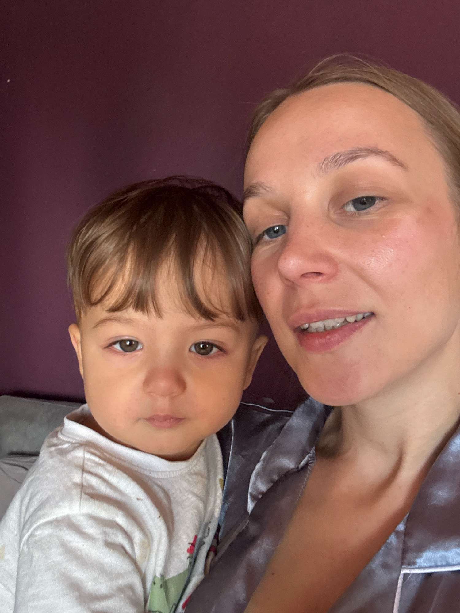 The image is a close-up selfie of a woman and a young child. The woman, positioned on the right side of the frame, has fair skin, light brown hair pulled back, and blue eyes. She is wearing a light-colored, possibly satin, pajama top. The child, on the left, appears to be a toddler with light brown hair and brown eyes. The child is wearing a white t-shirt with a graphic print. They are both looking towards the camera. The background is a dark purple wall. The scene appears to be indoors, likely in a residential setting. There is no indication of the location being in Überherrn, Germany.